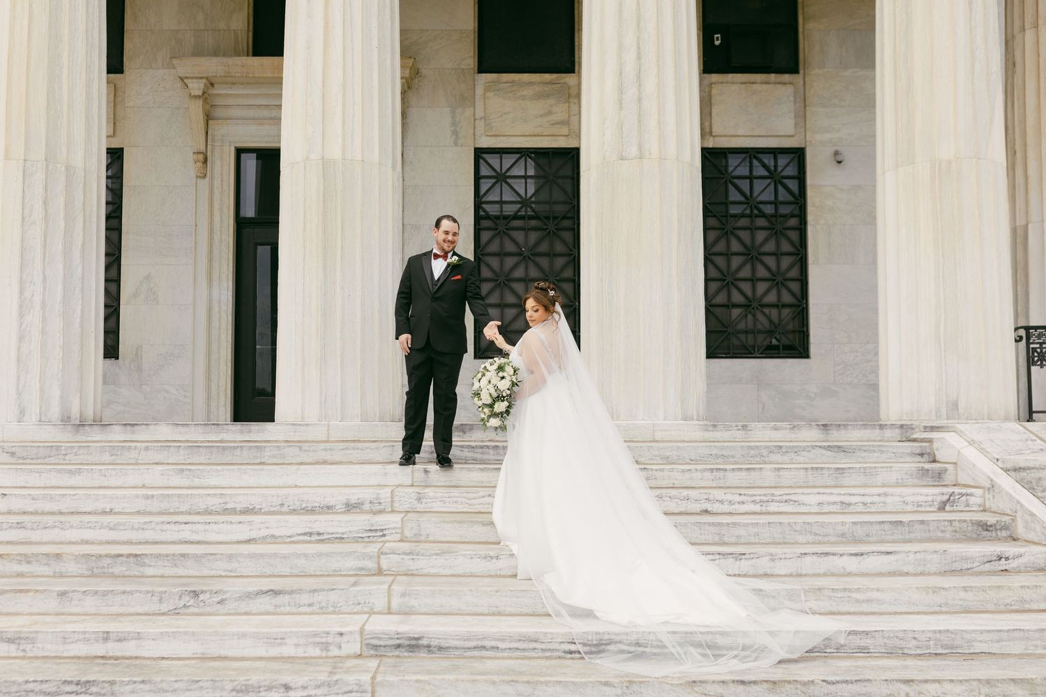 Bride and groom pose on grand white marble steps of a classical building on their wedding day.