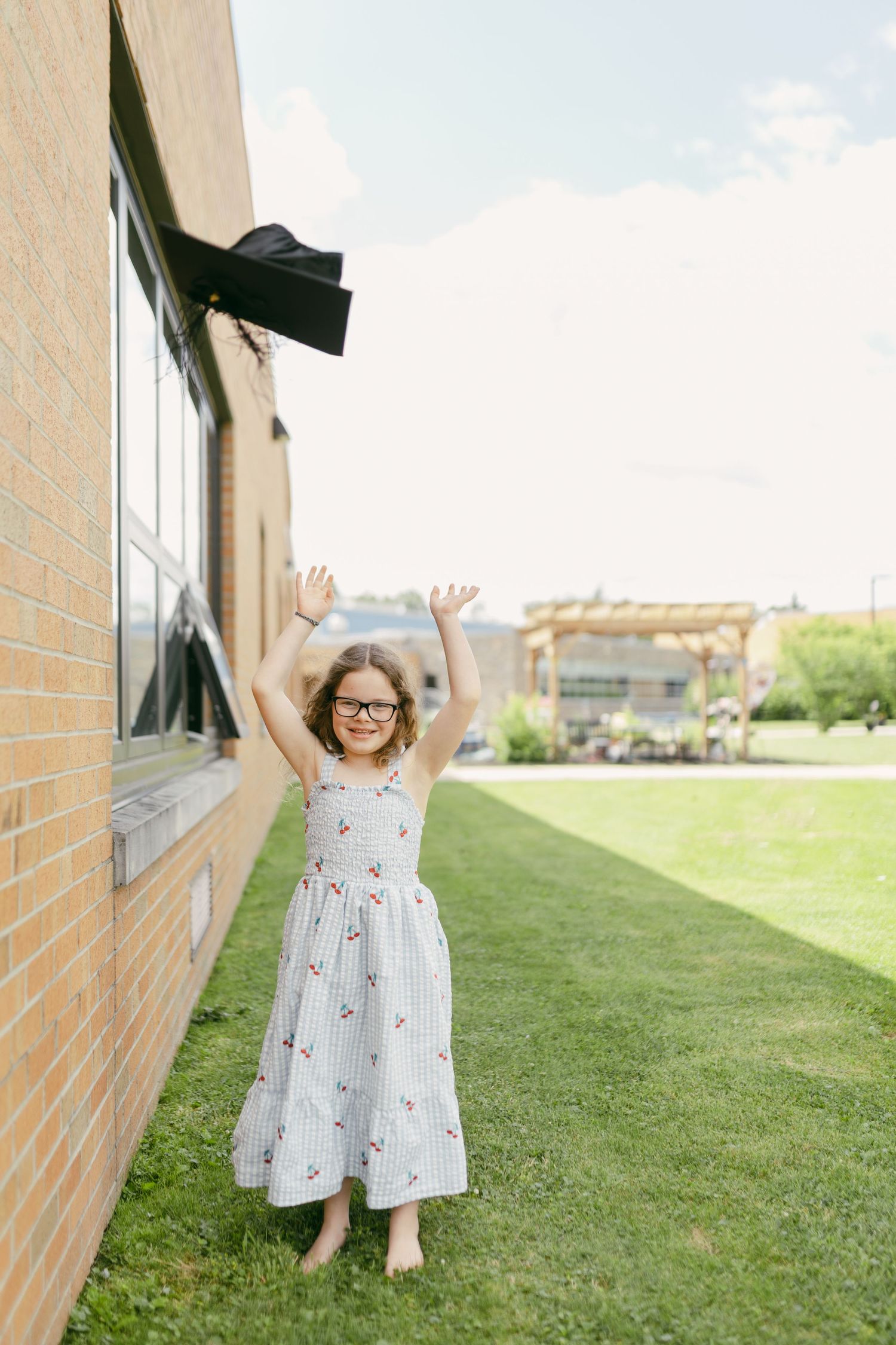 Young girl in floral dress and glasses tossing graduation cap in the air outdoors, celebrating academic achievement.
