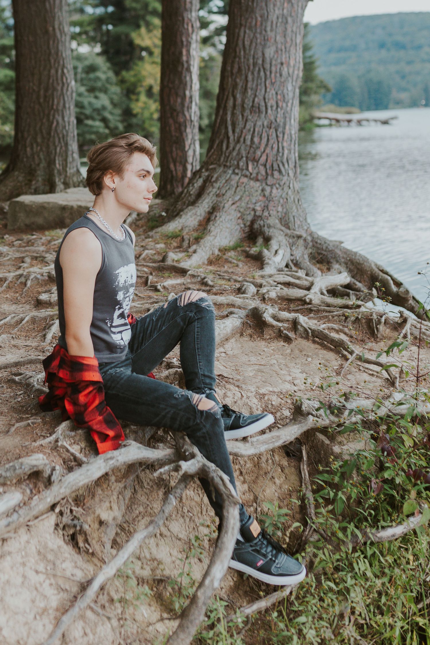 Young man in ripped jeans and tank top sitting on tree roots by a lake in a forest setting.
