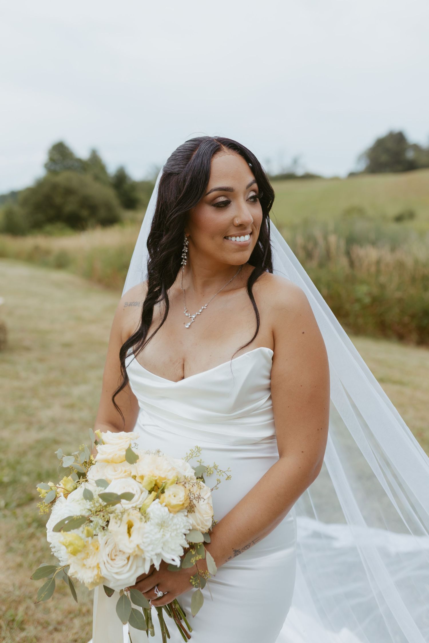 Smiling bride in white strapless gown and veil holds white floral bouquet outdoors in green field.