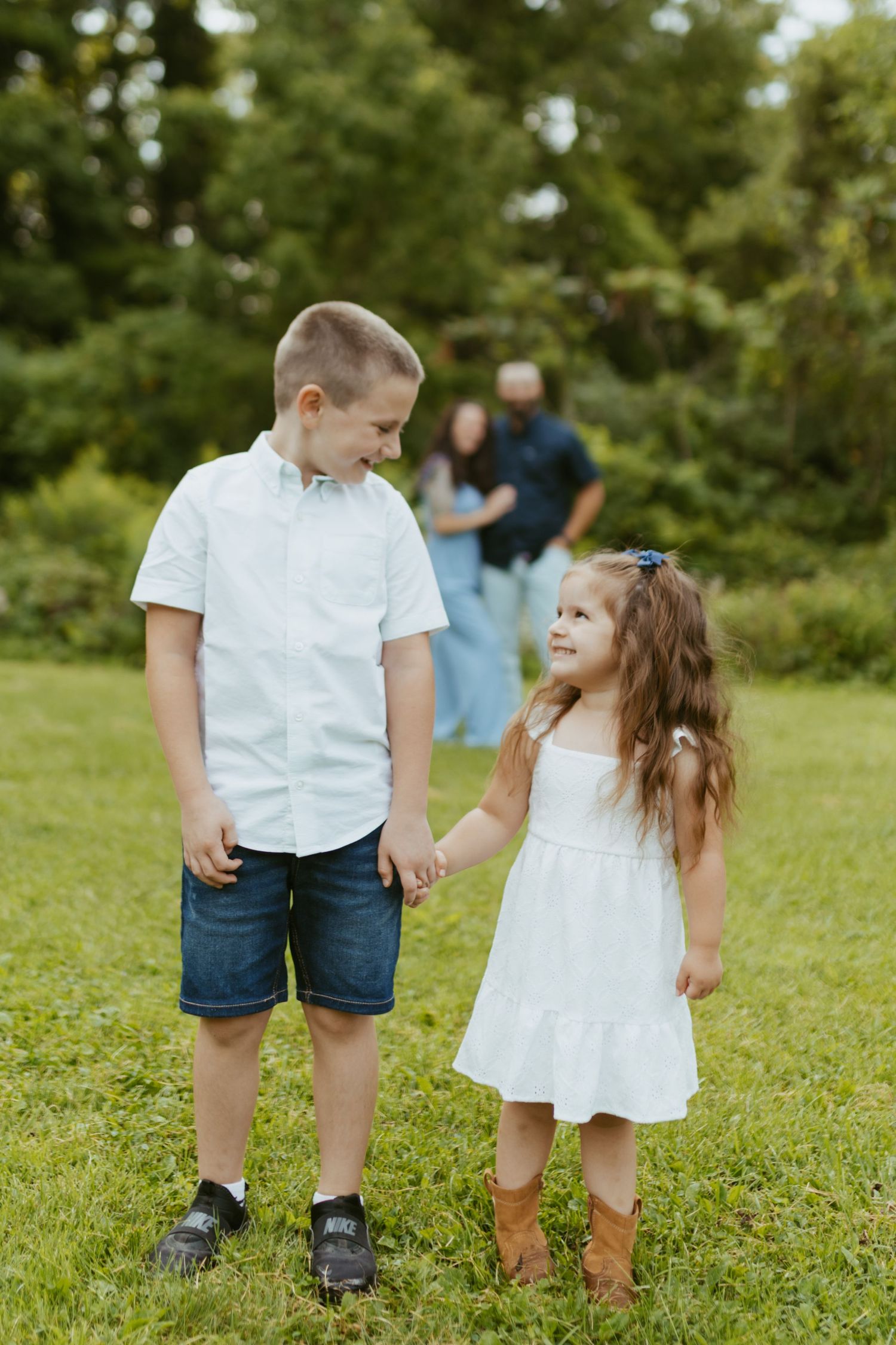 Two young siblings hold hands in a green park, boy in white shirt and girl in white dress with brown boots.
