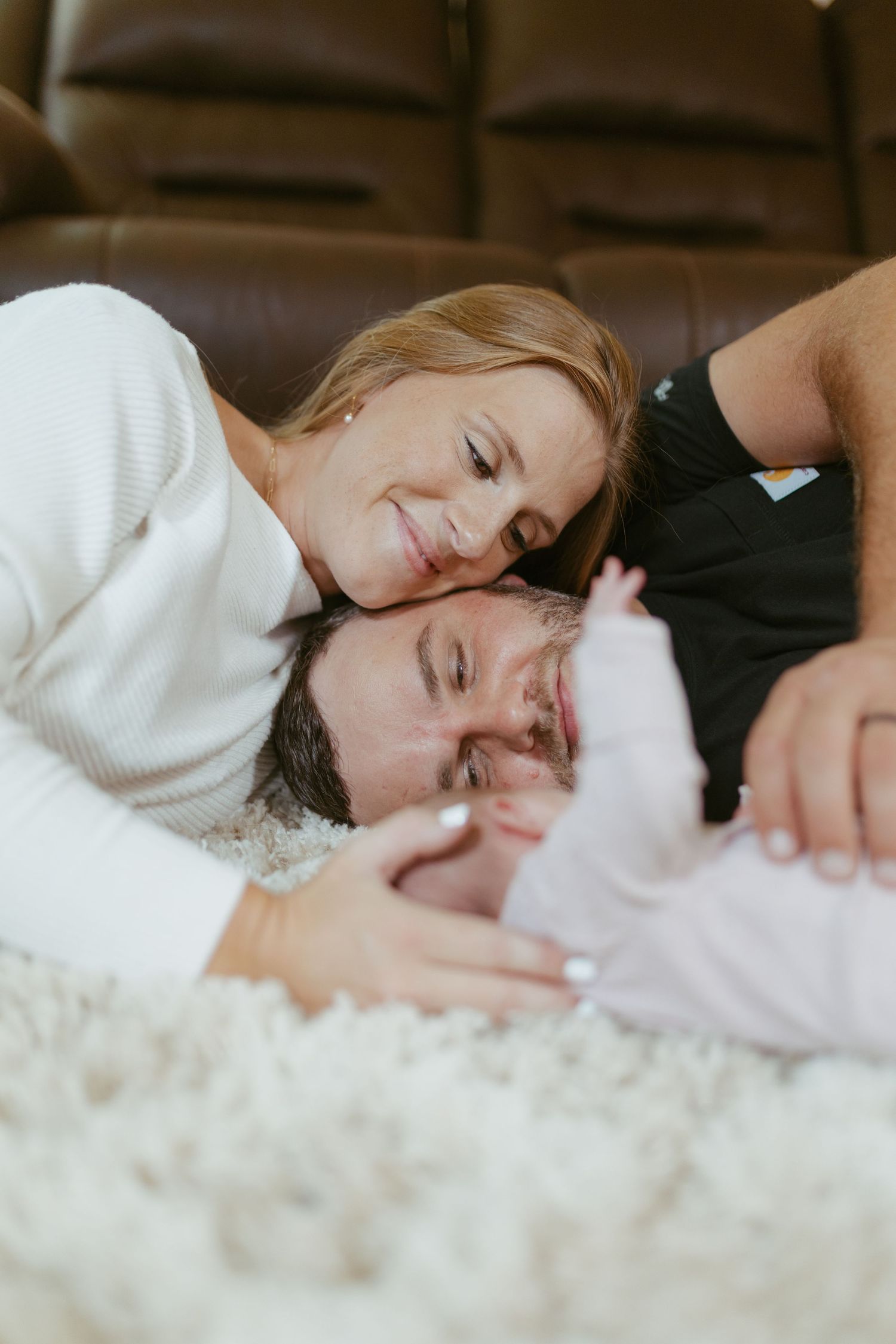 A smiling couple lying on a fluffy white rug, lovingly gazing at their newborn baby wrapped in pink.