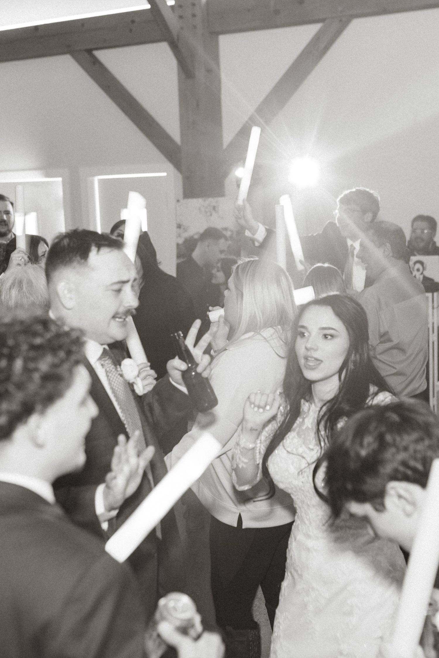 Black and white photo of wedding guests dancing with light-up foam sticks on the dance floor.