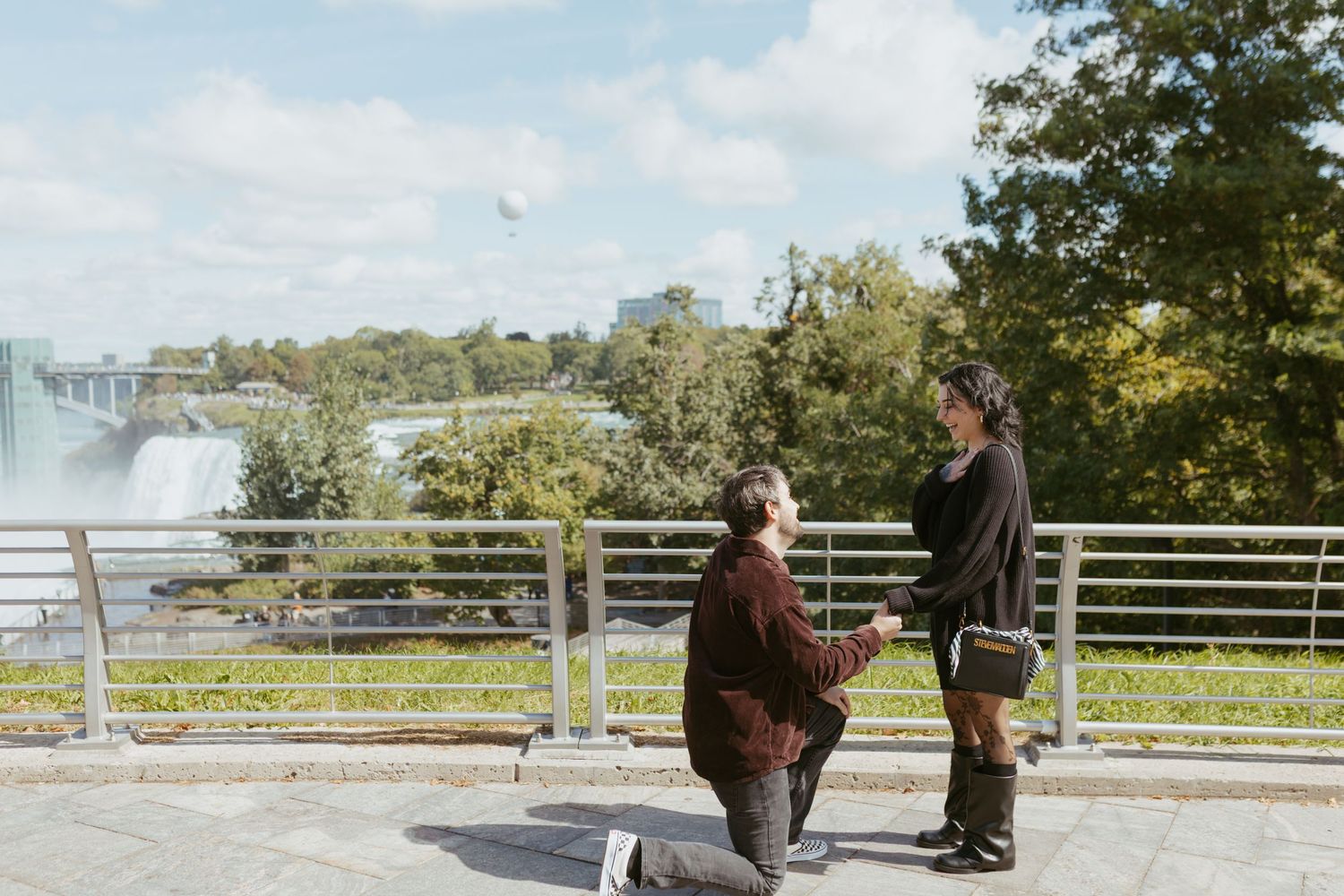 Man proposing on one knee to woman at Niagara Falls overlook on a sunny day.