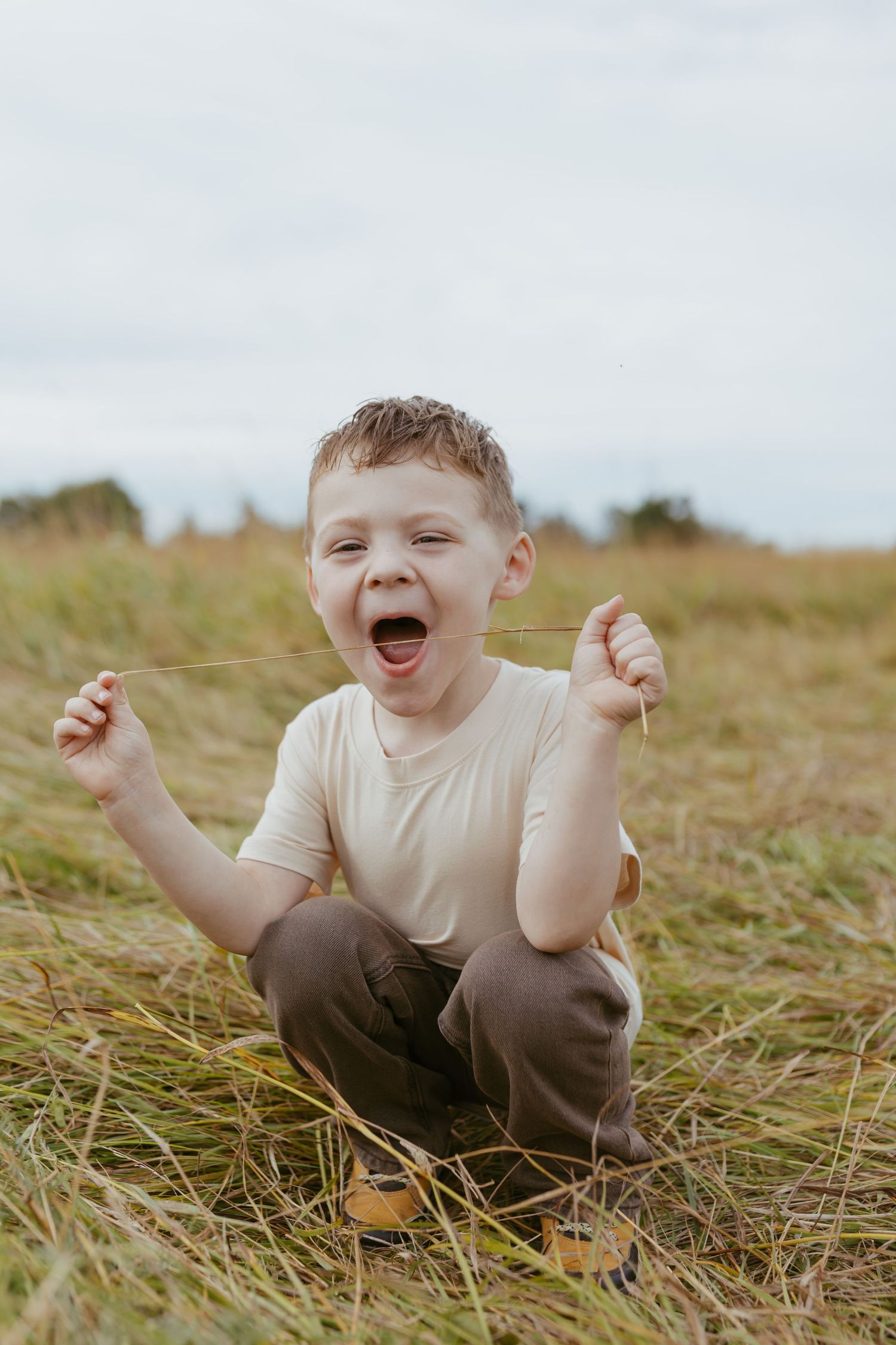 A joyful young boy sits in a grassy field, laughing and holding blades of grass in both hands.