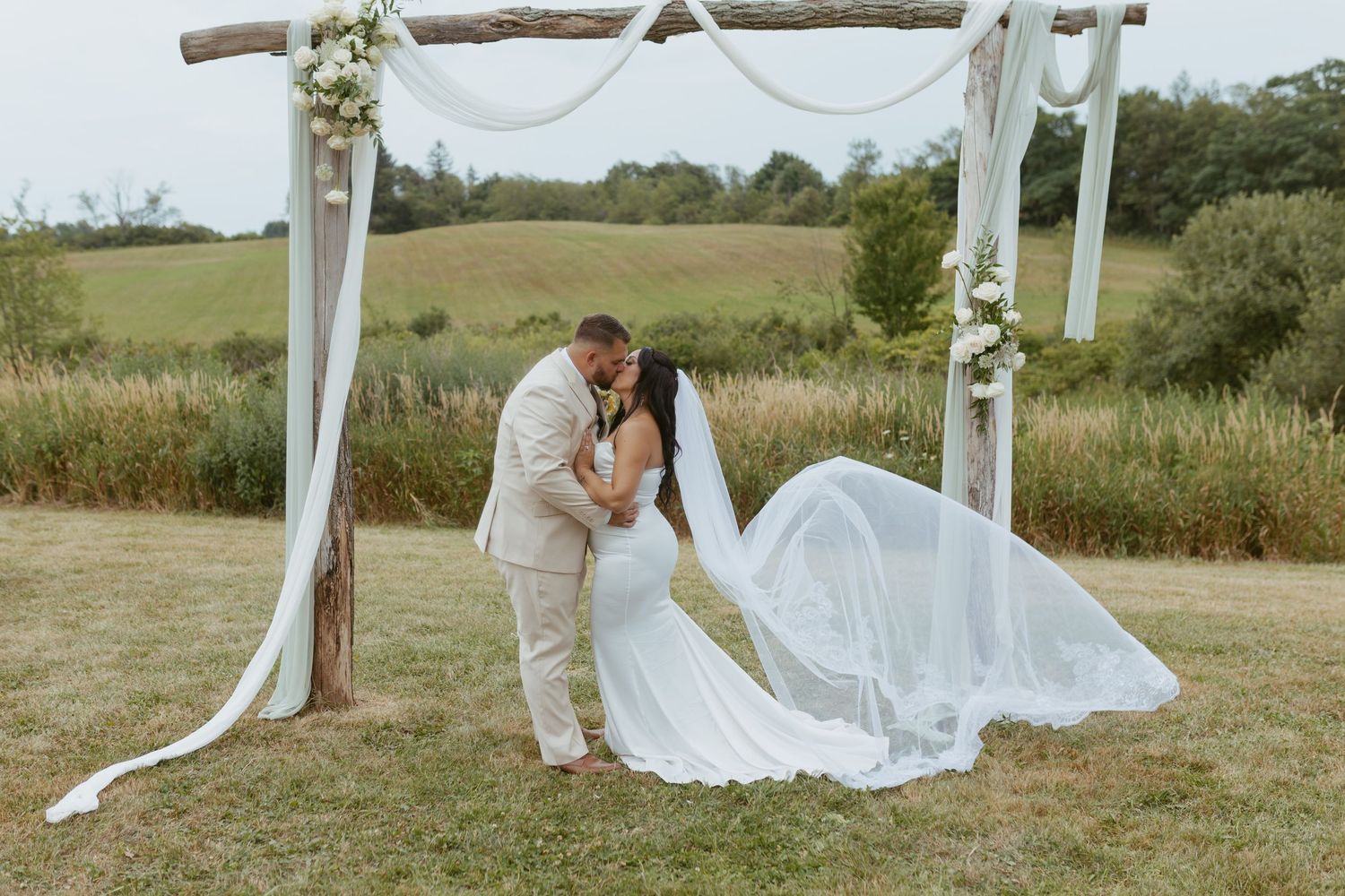 Bride and groom share a romantic kiss under a rustic wooden arch with white draping in a scenic outdoor wedding ceremony.