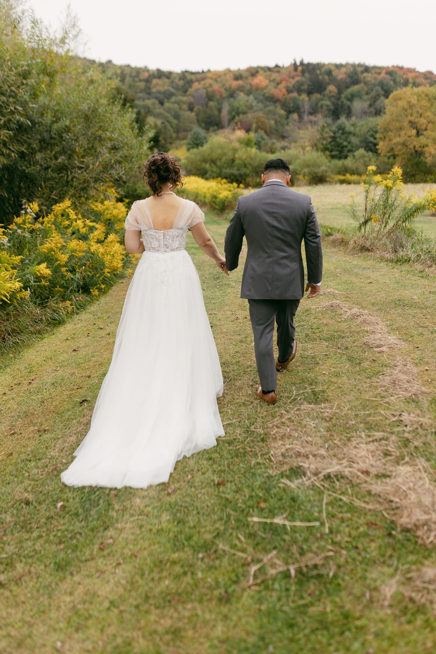 Newlyweds walk hand in hand through autumn field with golden wildflowers.