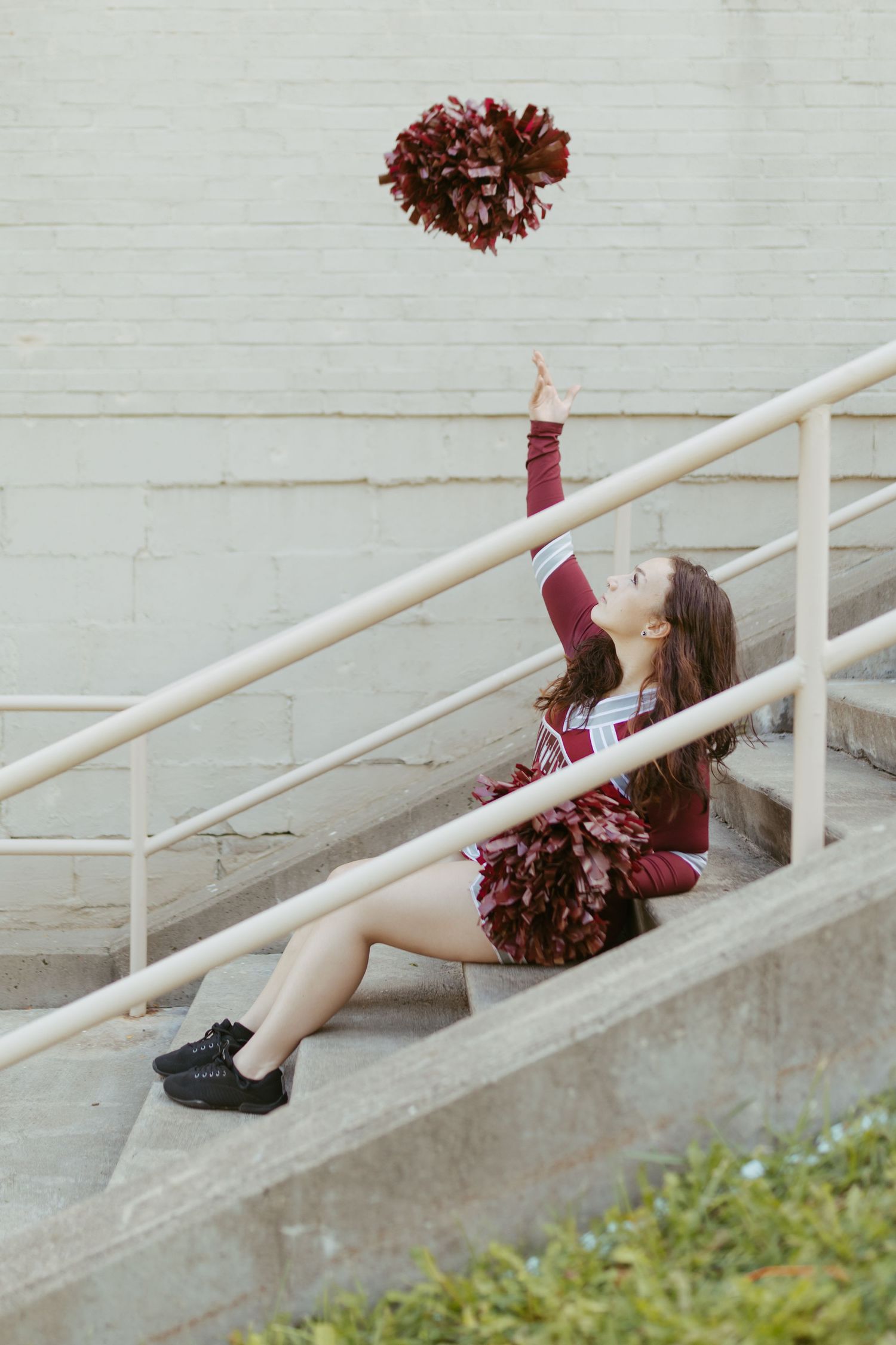 Cheerleader in maroon uniform sitting on stadium steps tossing a pom-pom in the air.