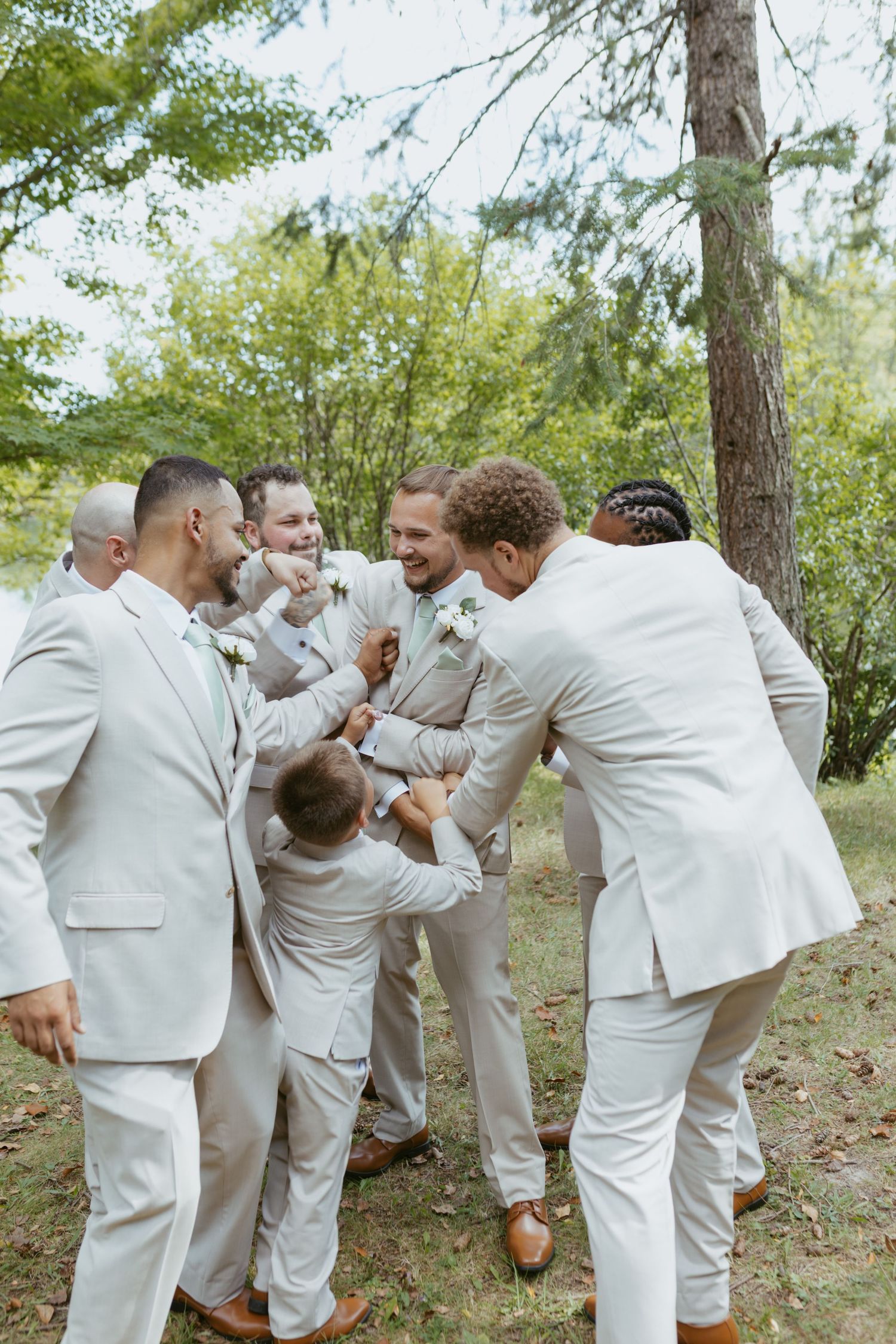 Groomsmen in light beige suits laughing and celebrating outdoors near trees on a wedding day.