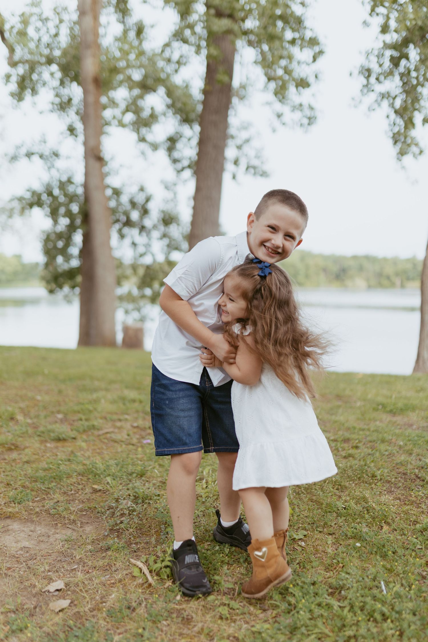 Two happy children hugging outdoors near a lake, boy in white shirt and girl in white dress smiling.