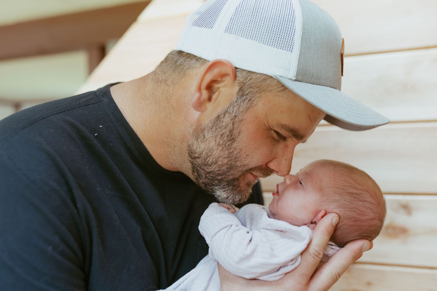 A bearded father in a gray cap tenderly kisses his newborn baby while cradling them gently in his hands.