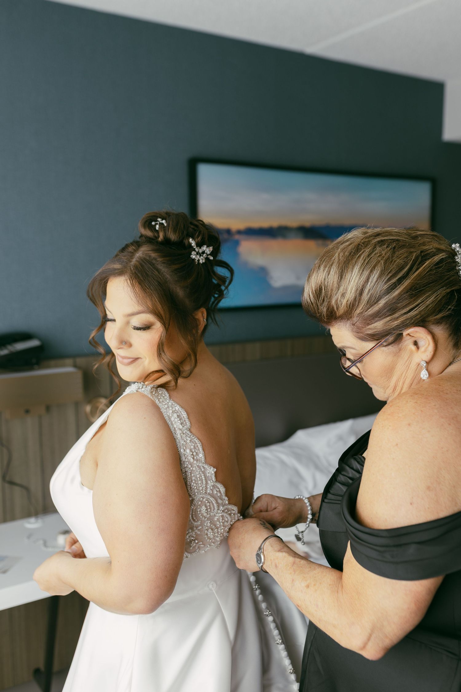 A bride in a white lace gown has her dress buttoned by an older woman in a black dress in a hotel room.