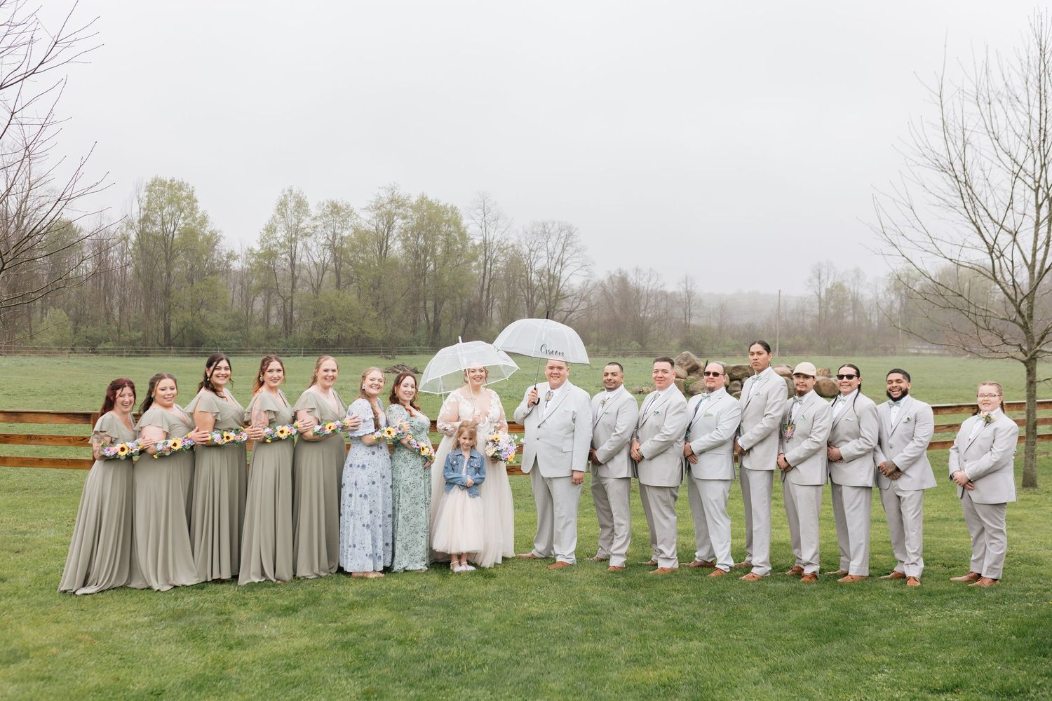 Wedding party poses outdoors on rainy day, bridesmaids in sage green gowns and groomsmen in tan suits with bride holding umbrella.