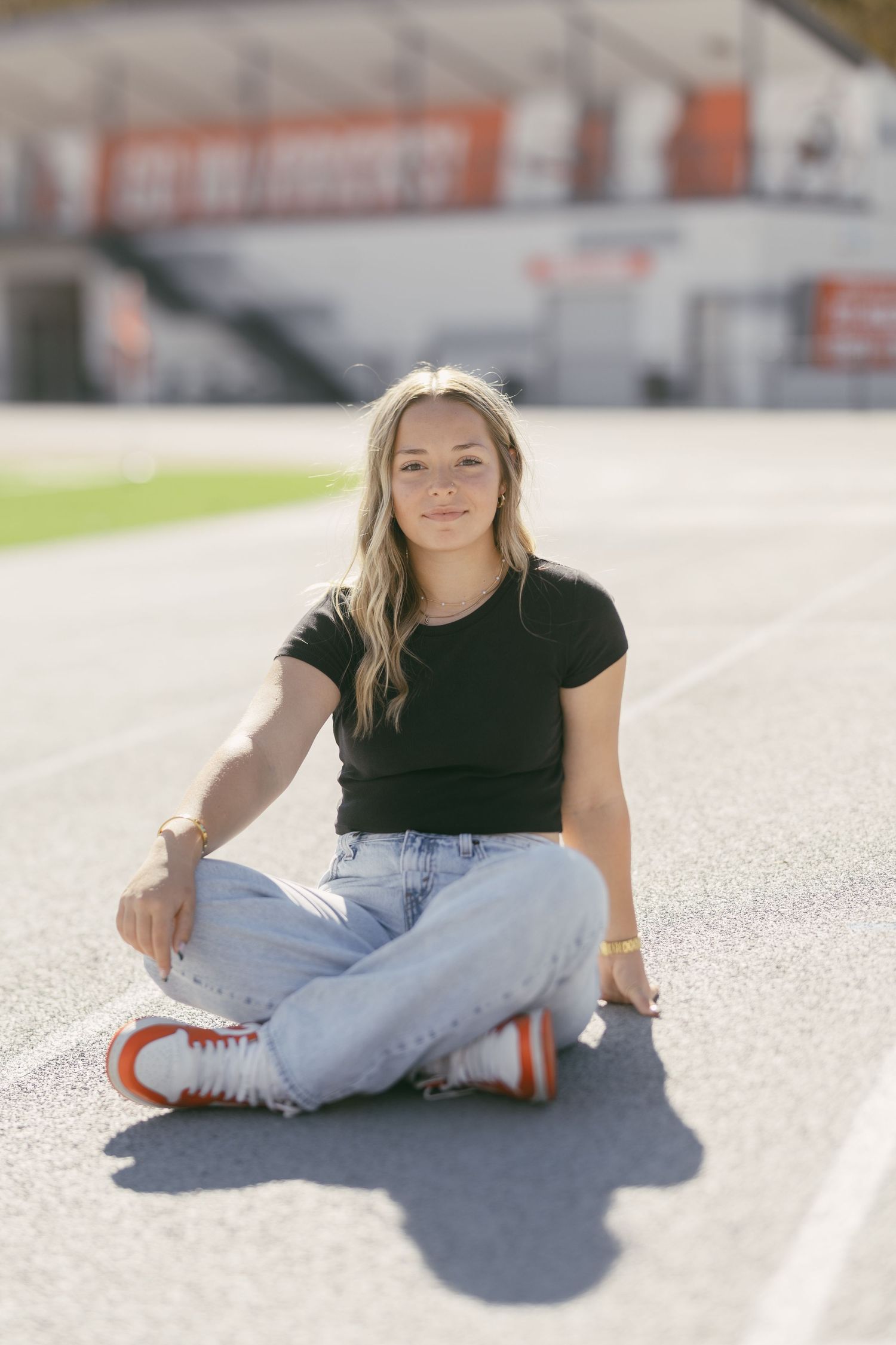 Young woman in black tee and jeans sits cross-legged on a stadium track, wearing orange sneakers, smiling at camera.
