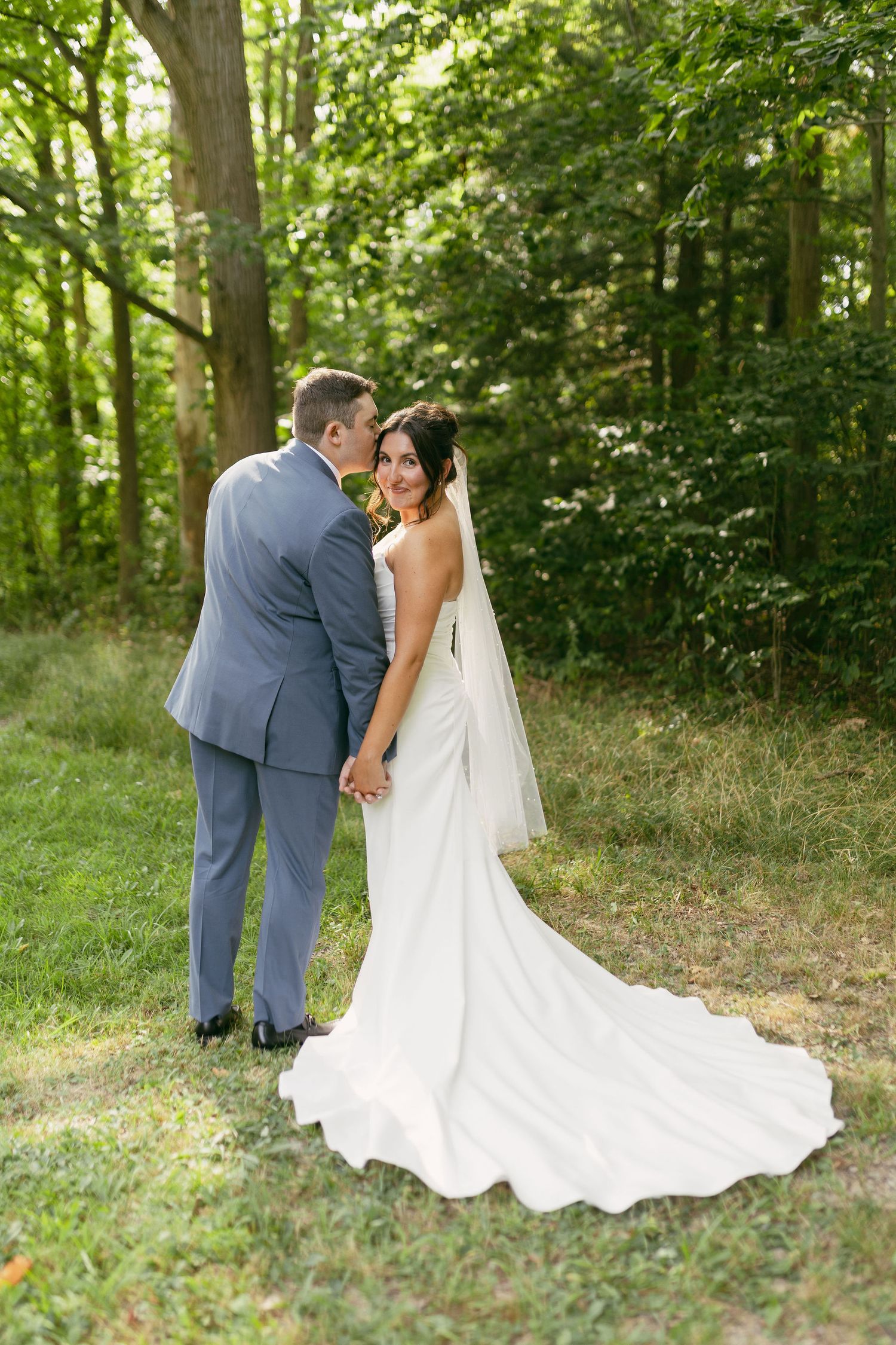 Couple poses in elegant wedding attire against a backdrop of lush green forest.