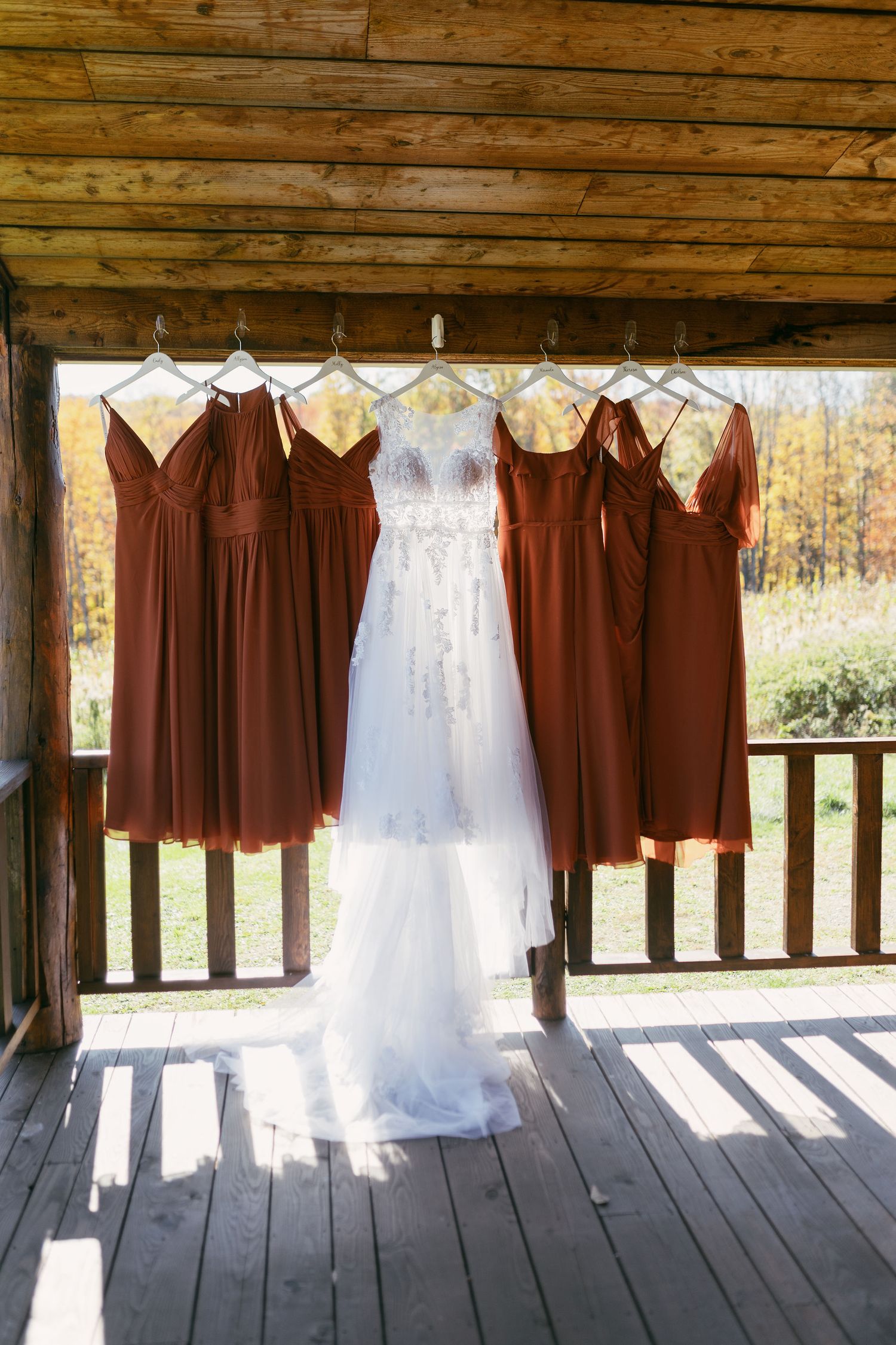 Orange bridesmaid dresses and white wedding gown hanging in a row on rustic wooden porch.