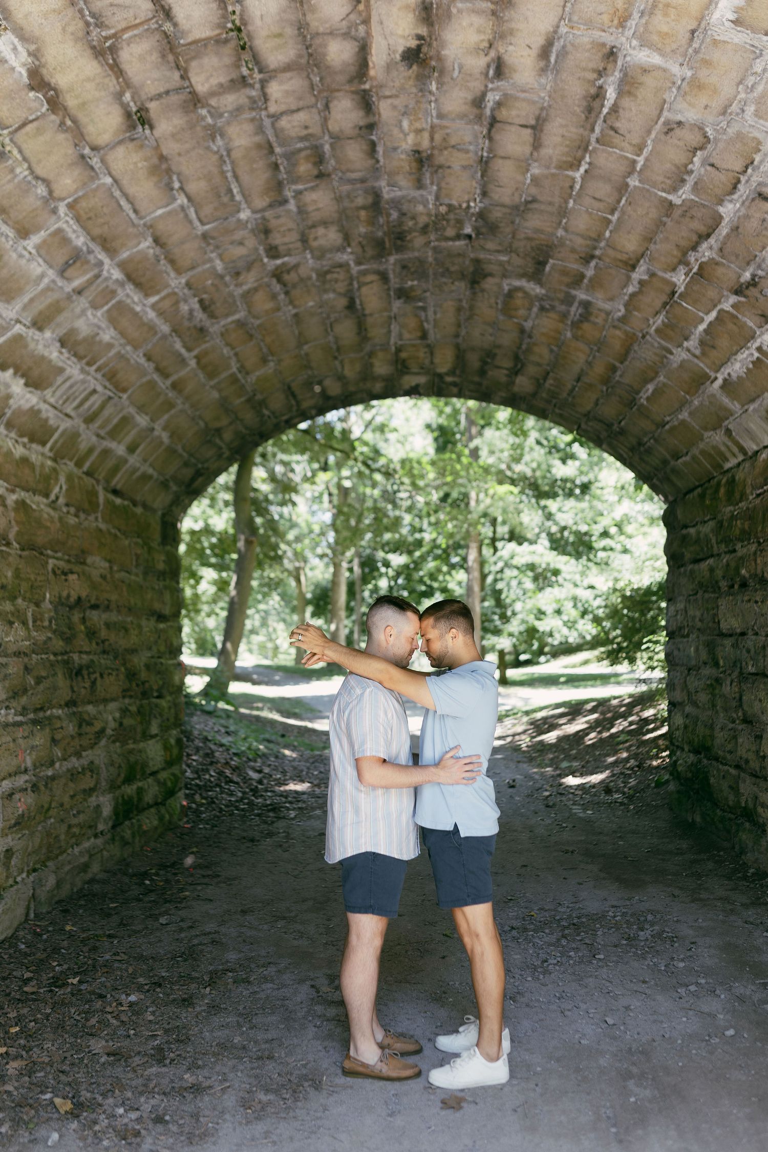 Two people share an intimate moment under a brick archway tunnel in a park.