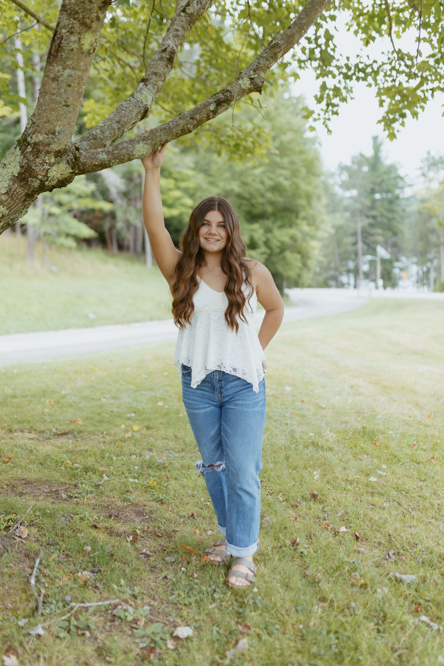 Young woman in white tank top and ripped jeans poses under a tree branch on a green lawn.