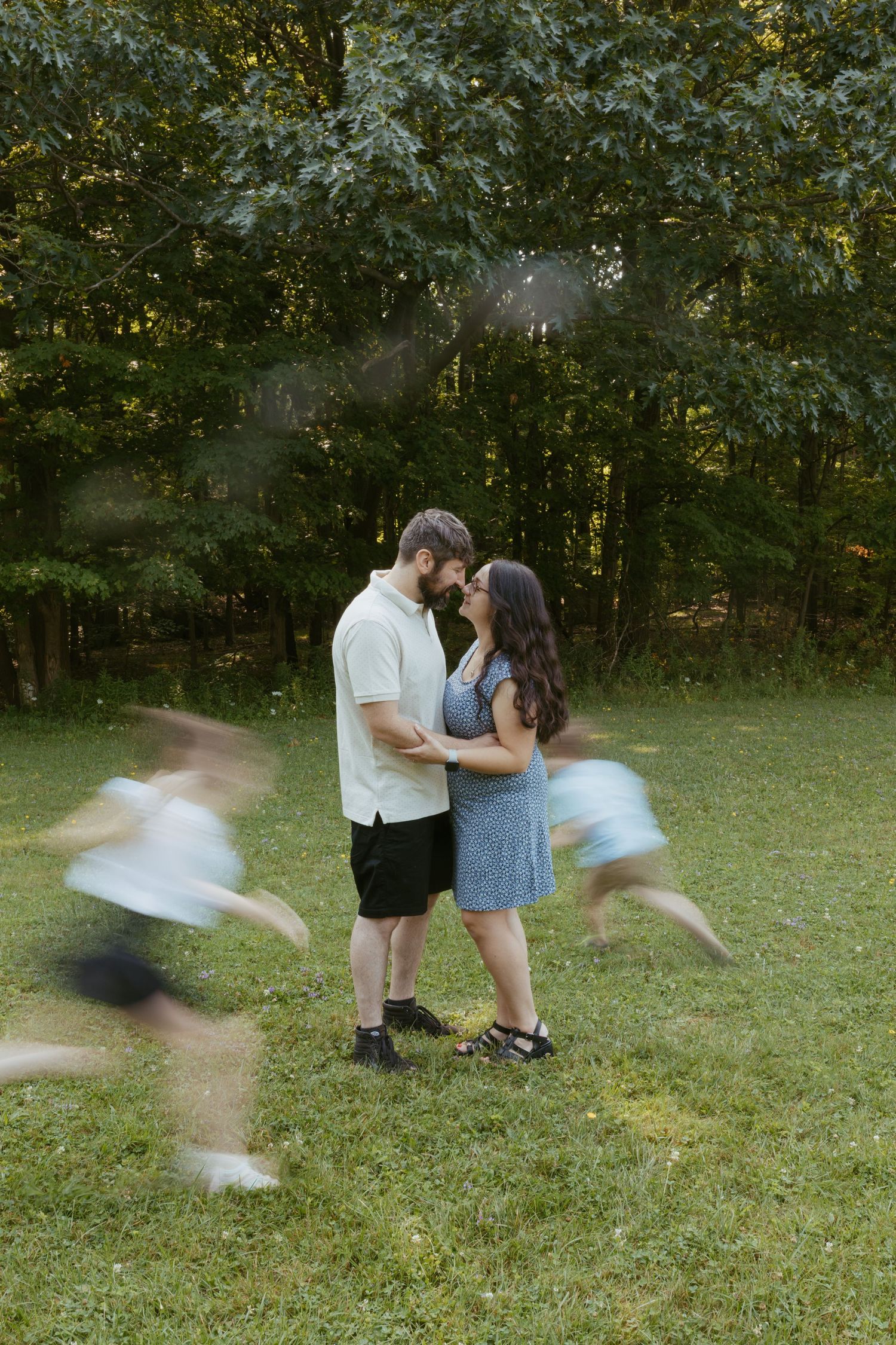Couple shares romantic moment in green park while blurred children run around them, captured with slow shutter speed.