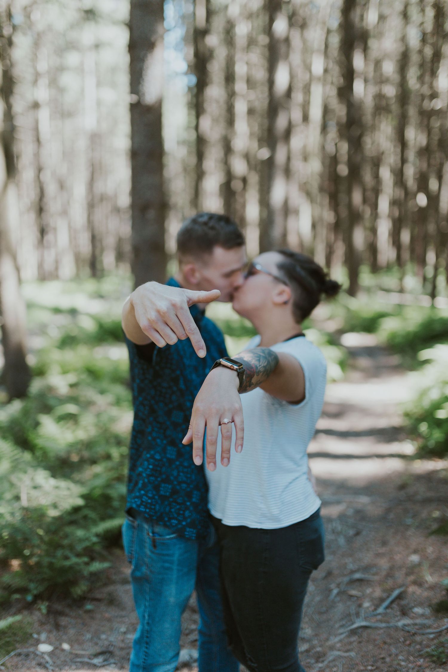 Couple kissing in forest while showing off engagement ring on woodland path.
