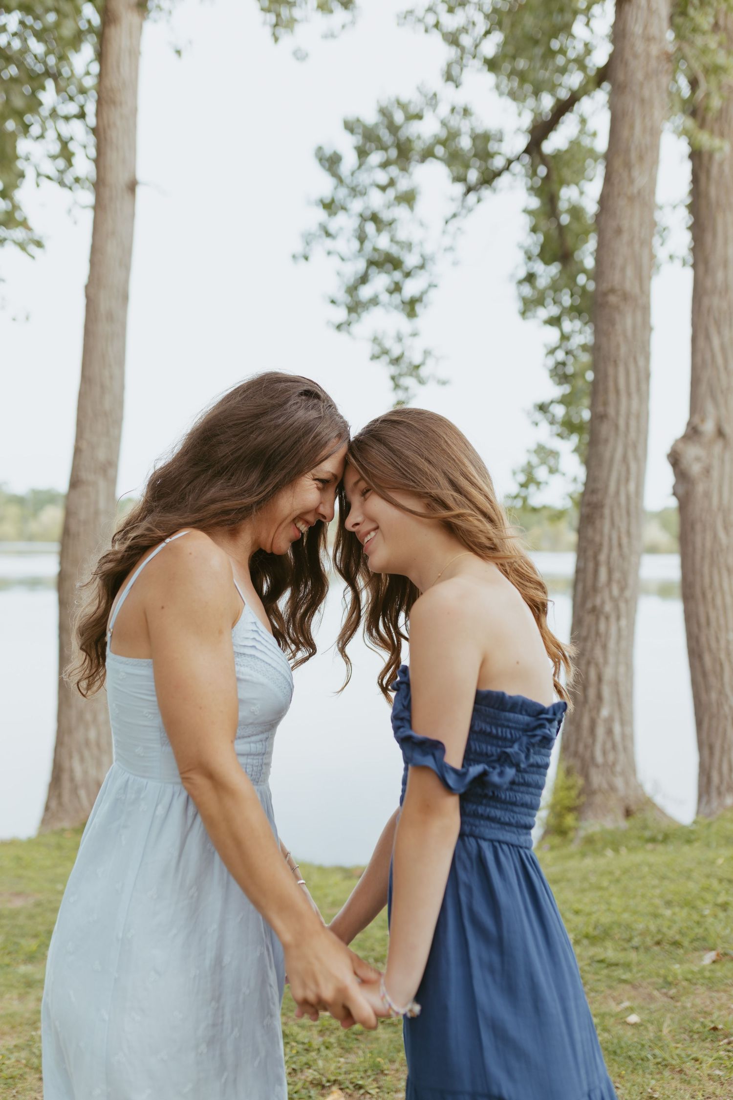 Two young women in summer dresses hold hands and touch foreheads, smiling near a lakeside with tall trees.