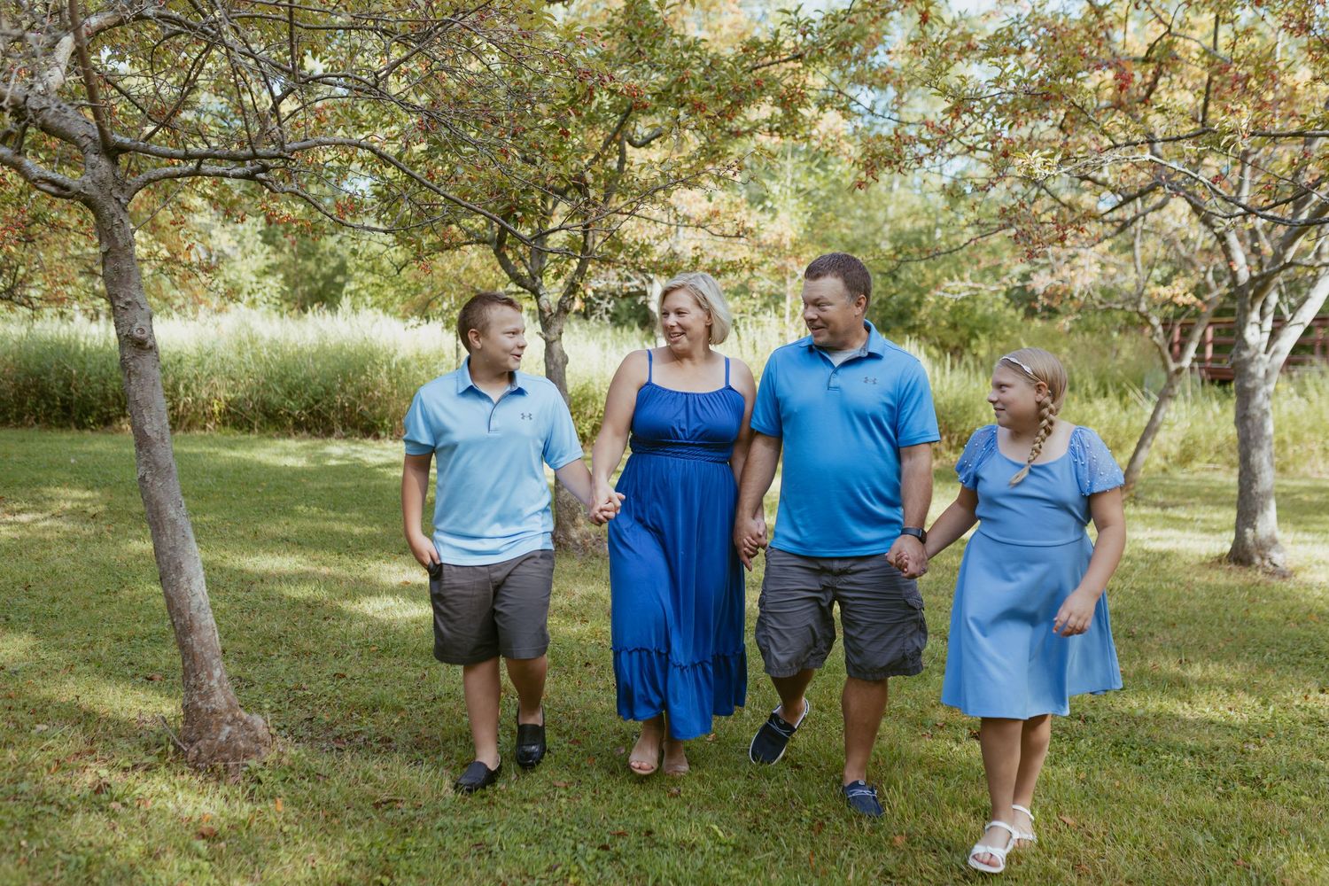 Family of four walks hand-in-hand through a lush green park, wearing coordinating blue outfits, smiling joyfully.