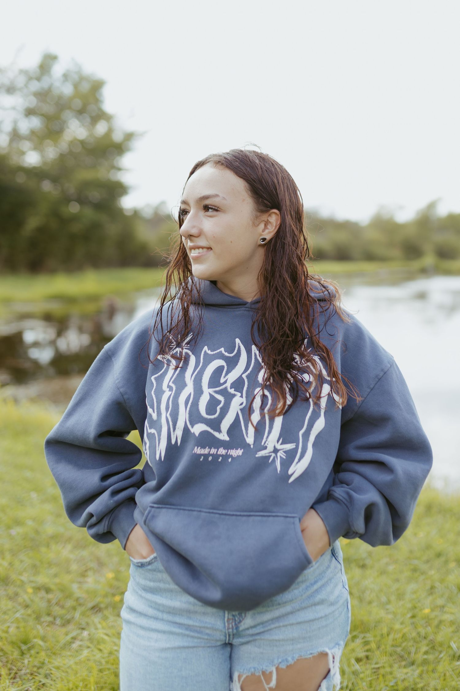 Young woman in blue graphic hoodie and ripped jeans smiling outdoors near a pond on a cloudy day.