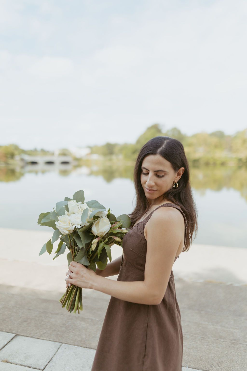 Woman in brown dress holding white flower bouquet by a serene lakeside, gazing down at flowers.