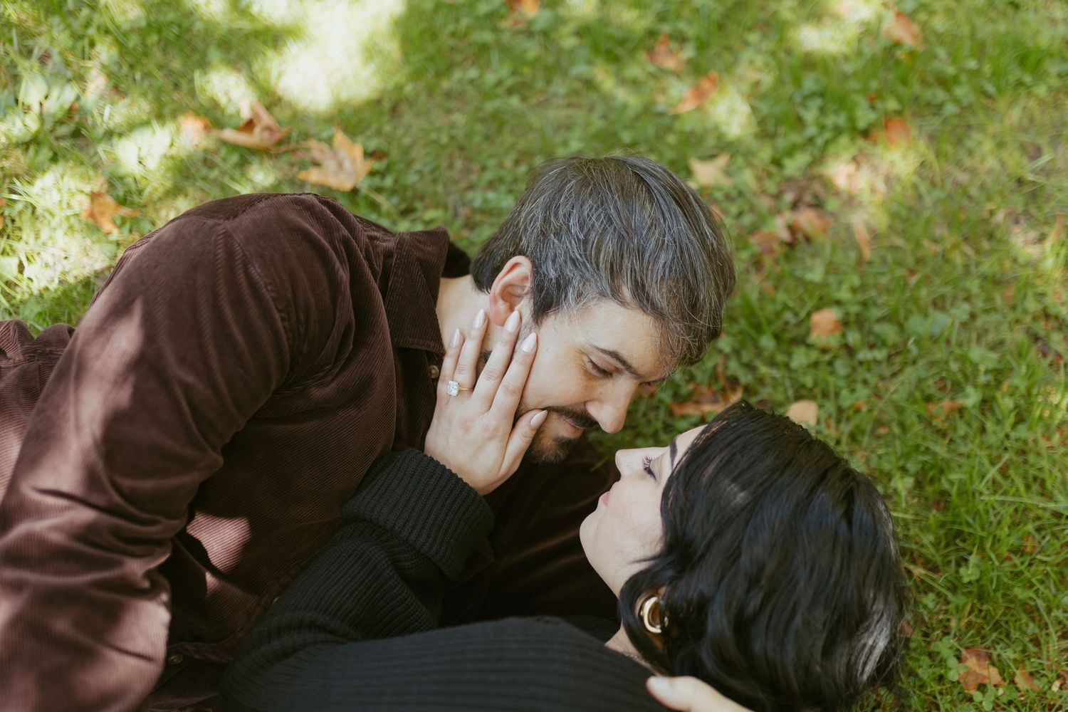 Romantic couple lying on green grass sharing a tender kiss outdoors in autumn park setting.