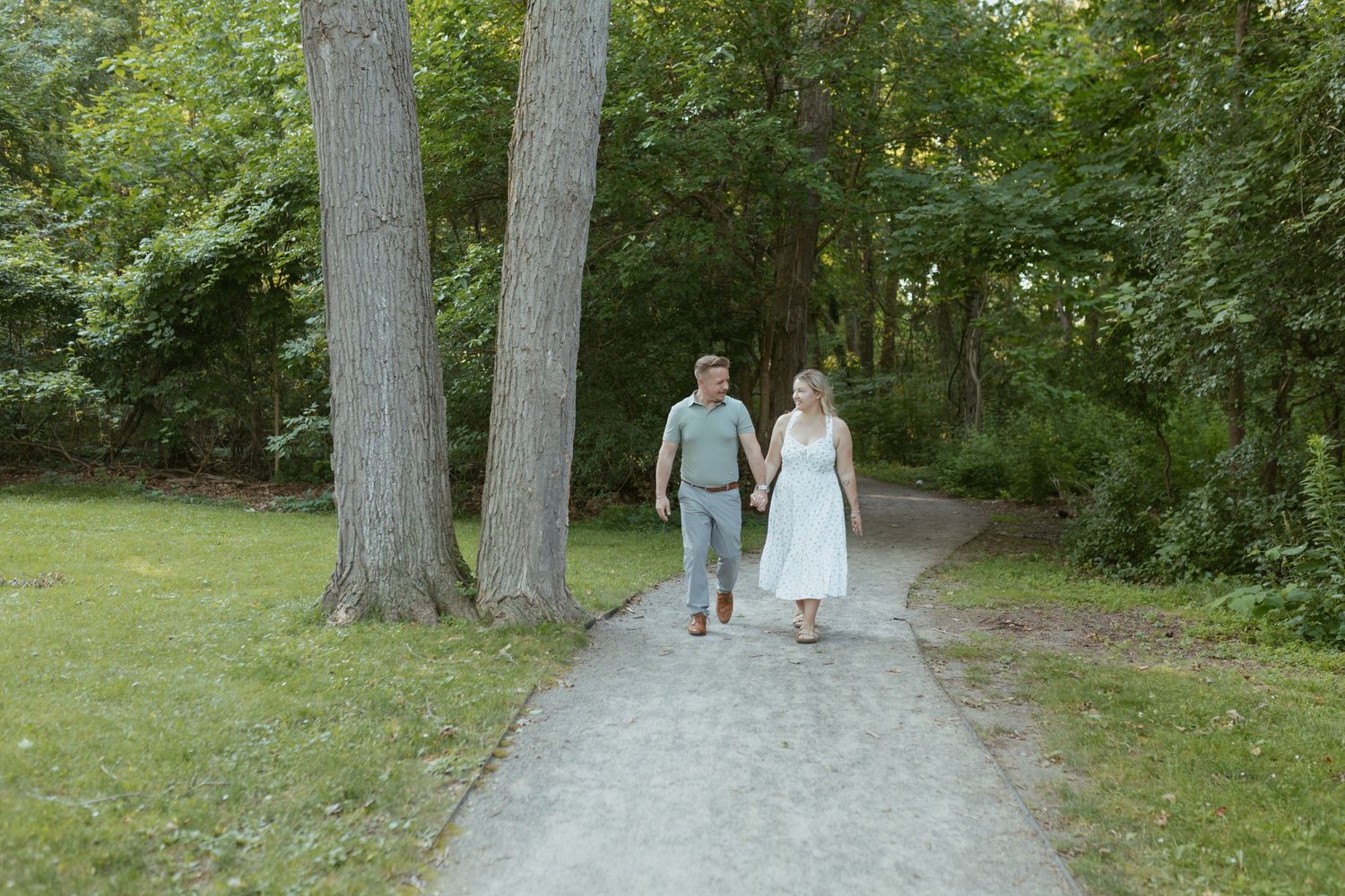 Couple holding hands walking together on a winding path through a lush green park surrounded by tall trees.