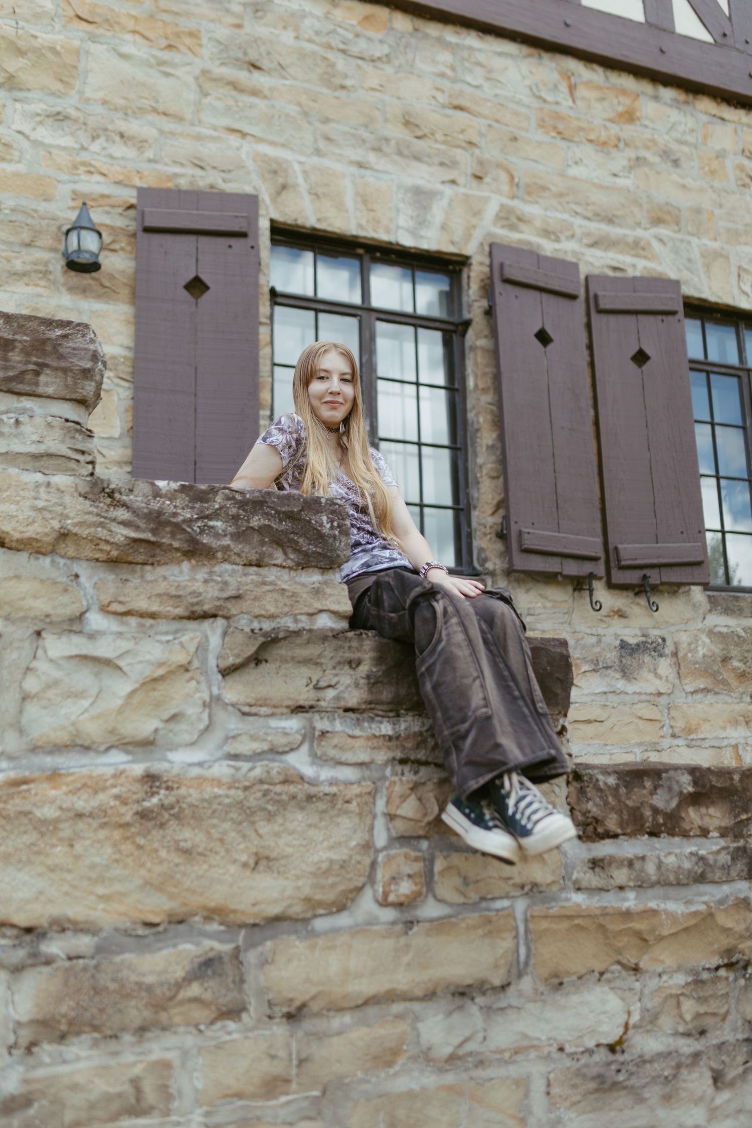 Young woman with long blonde hair sits on stone steps of a rustic building with brown shuttered windows.
