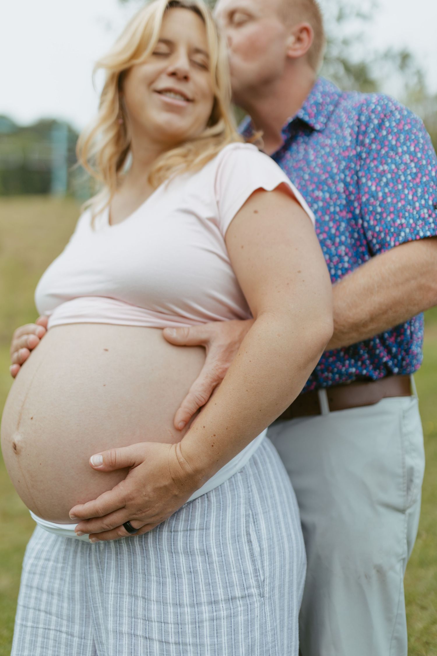 A pregnant woman smiles as her partner kisses her head and cradles her baby bump outdoors.