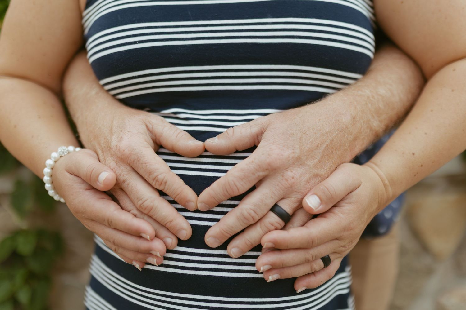 Pregnant woman in striped dress with partner's hands forming heart shape on baby bump.