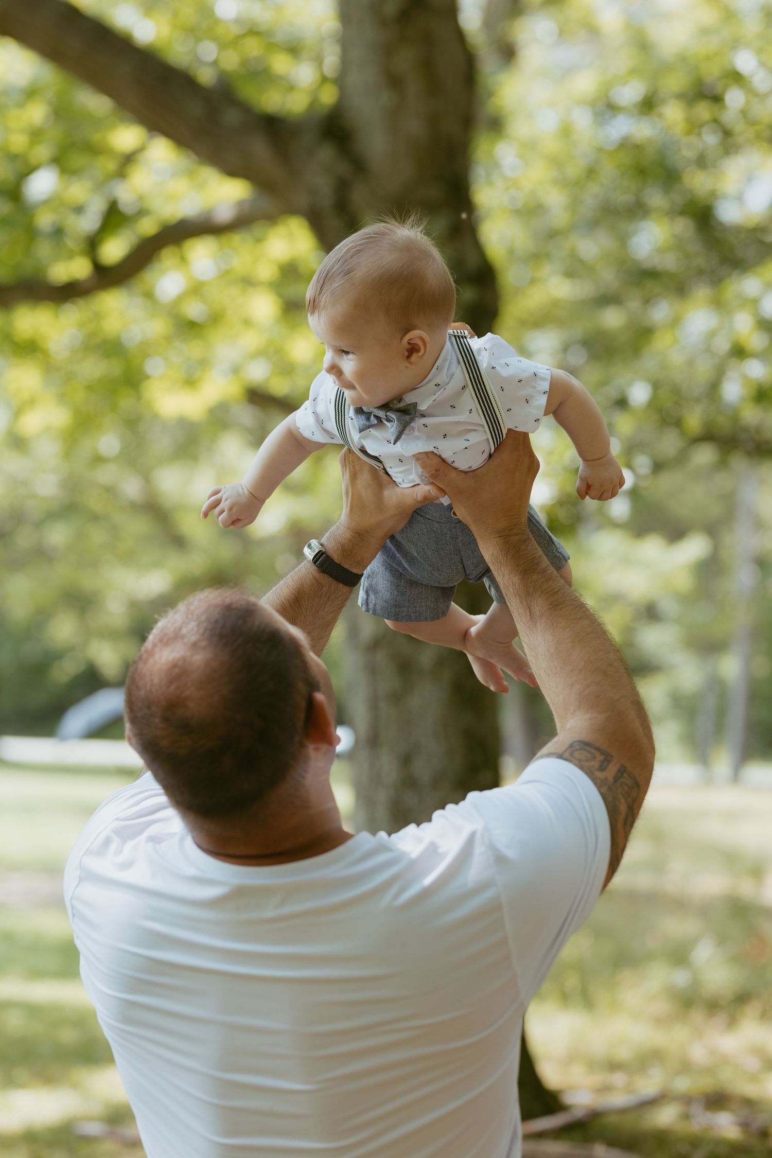 Father joyfully lifting baby in the air outdoors in a sunny park with lush green trees in the background.