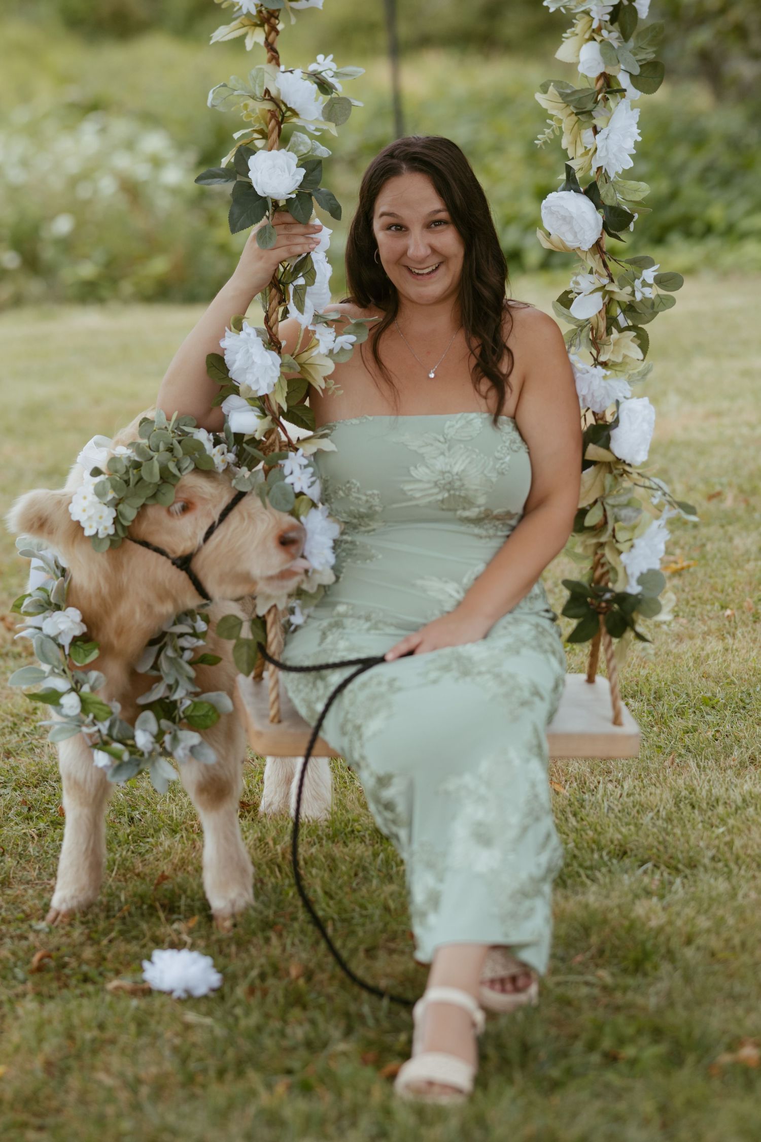 Woman in sage green dress on floral swing with baby goat wearing flower crown outdoors.
