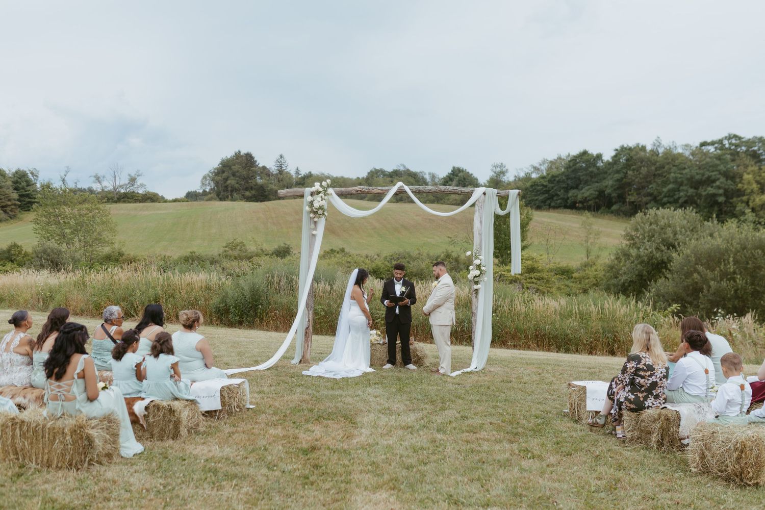 Outdoor wedding ceremony with couple at floral arch, guests seated on hay bales in a scenic green countryside setting.
