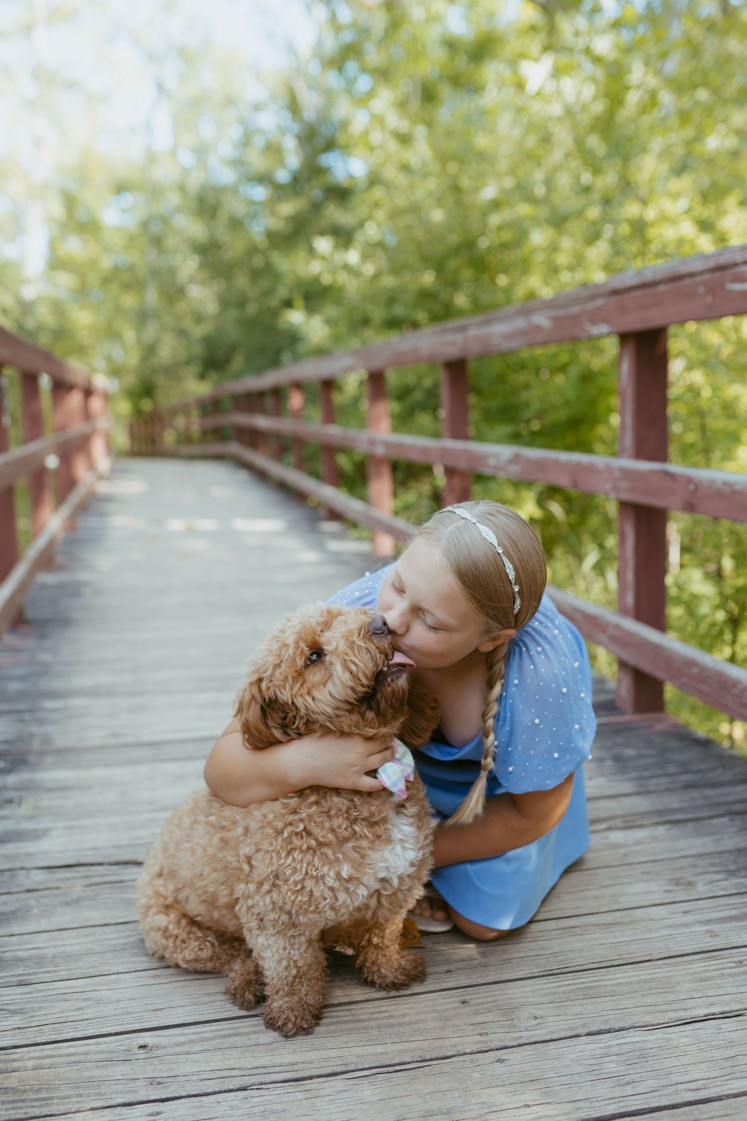 Young girl in blue dress hugging and kissing her fluffy golden doodle dog on a wooden bridge outdoors.