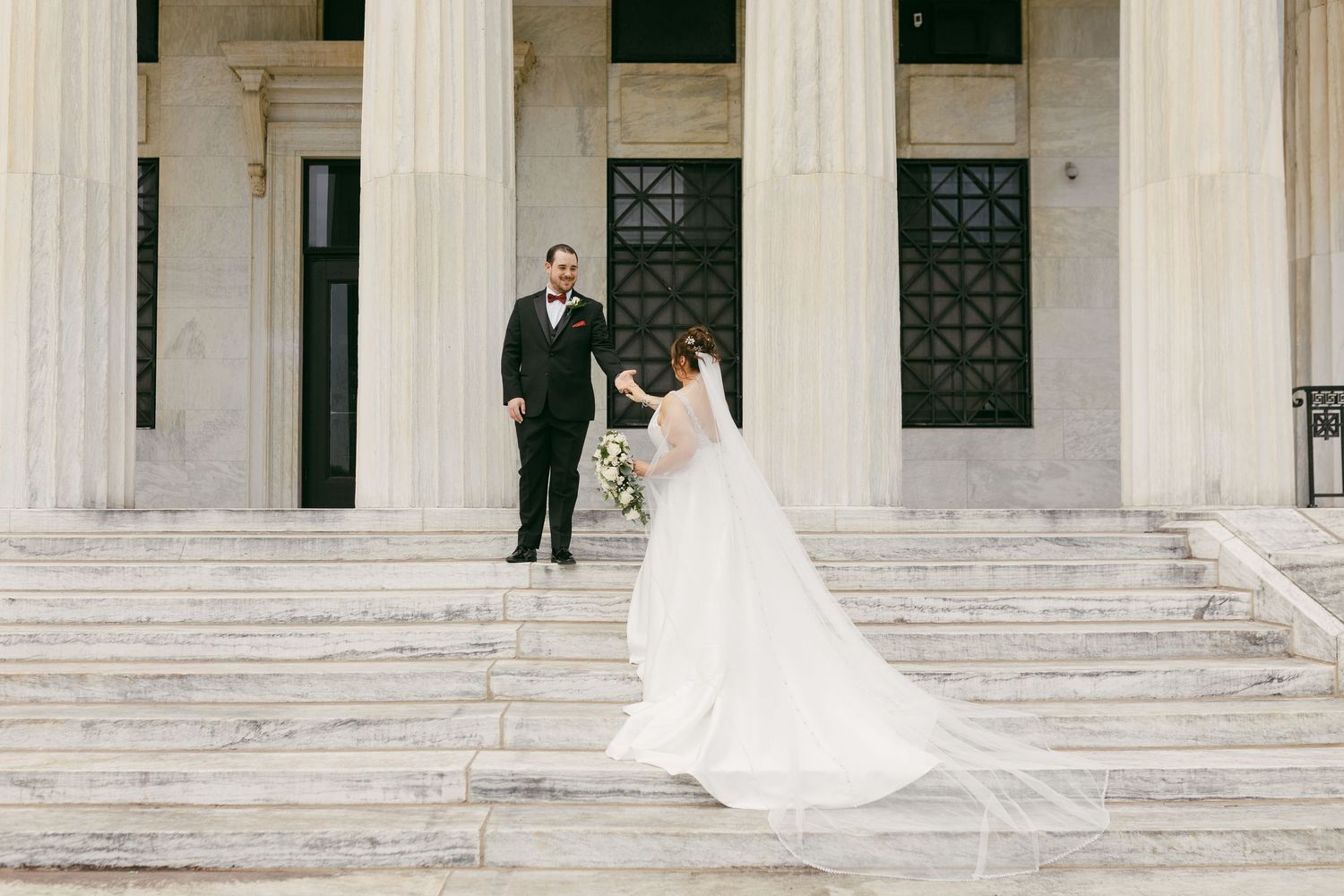 Bride in flowing white gown and veil walks toward groom in black tuxedo on grand marble steps of elegant columned building.