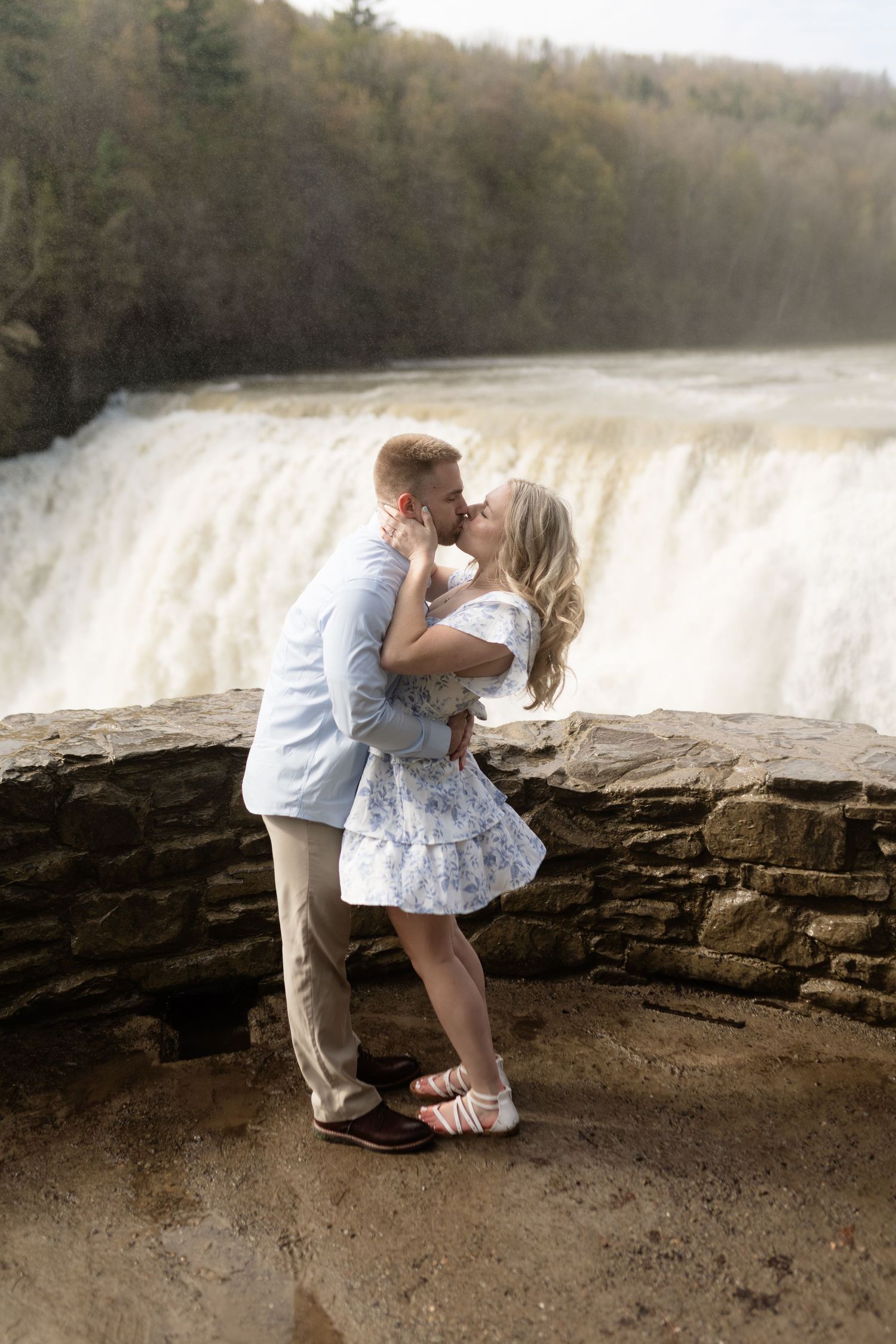 Couple sharing a romantic kiss in front of a rushing waterfall on a stone ledge, woman in white floral dress.