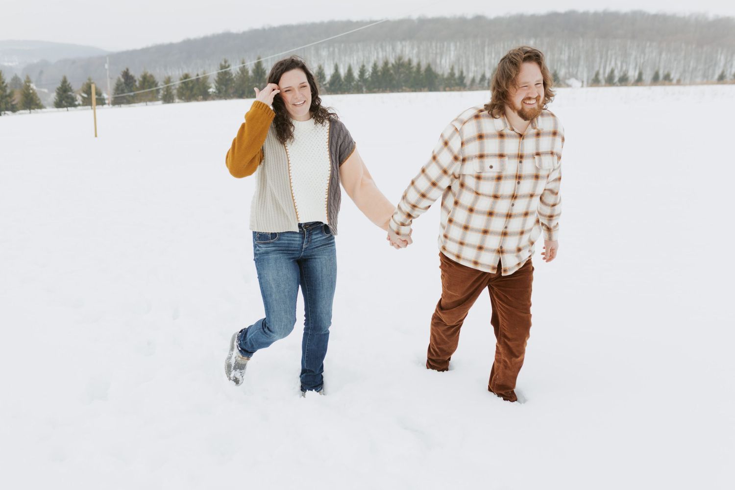 Couple holding hands and laughing while walking through deep snow in a winter landscape.