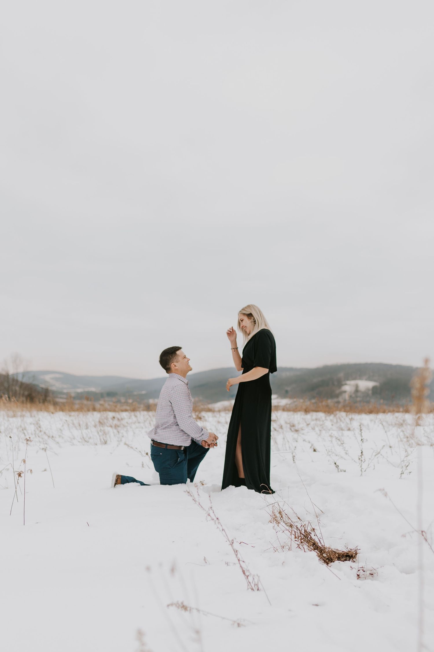 Man proposing to woman in snowy winter field, both dressed elegantly, with snow-covered hills in the background.