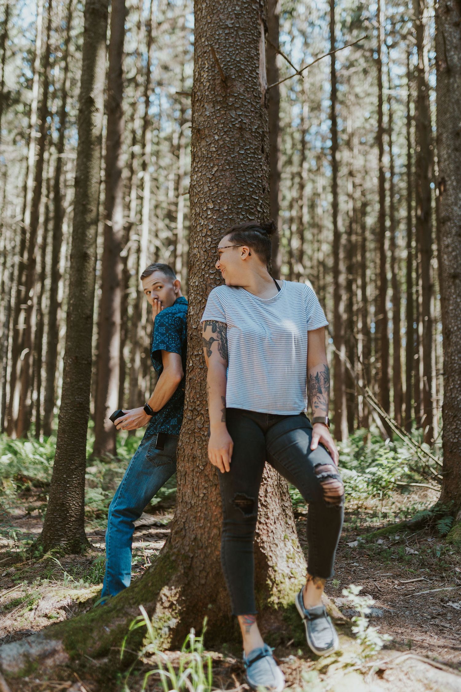 Two people leaning against a tree trunk in a sunlit forest, one with tattoos wearing a white tee and ripped jeans.