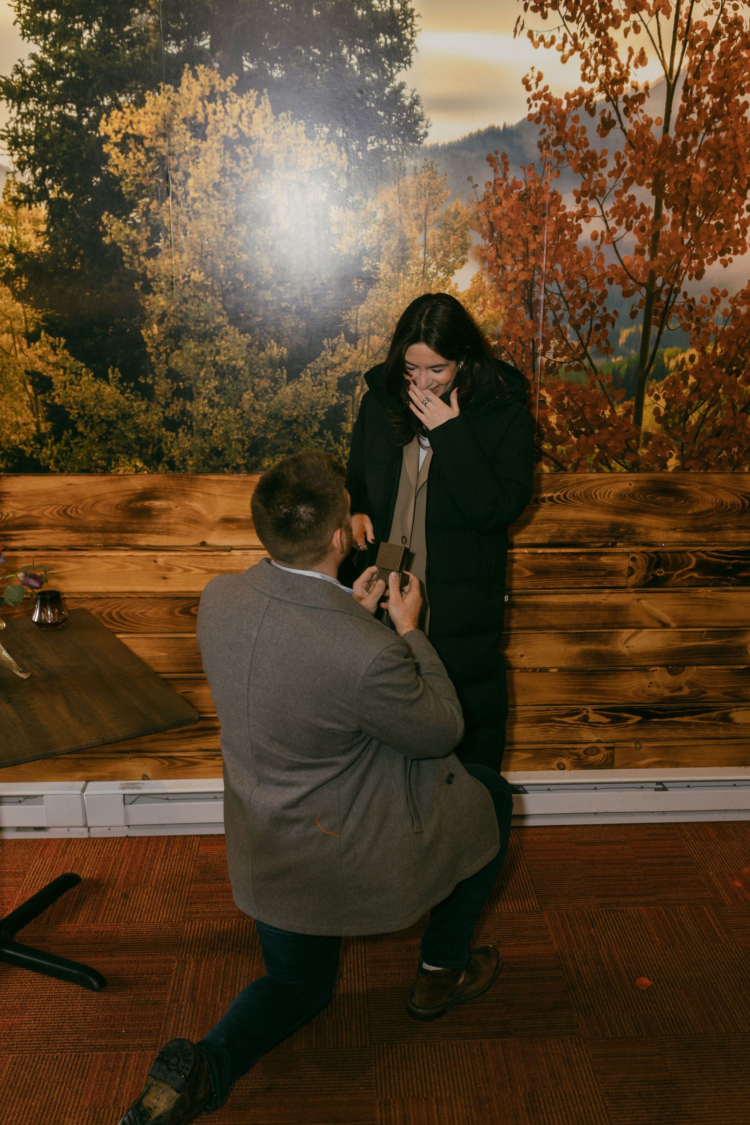 Man proposing on one knee to surprised woman covering her mouth in an autumn-themed indoor setting.