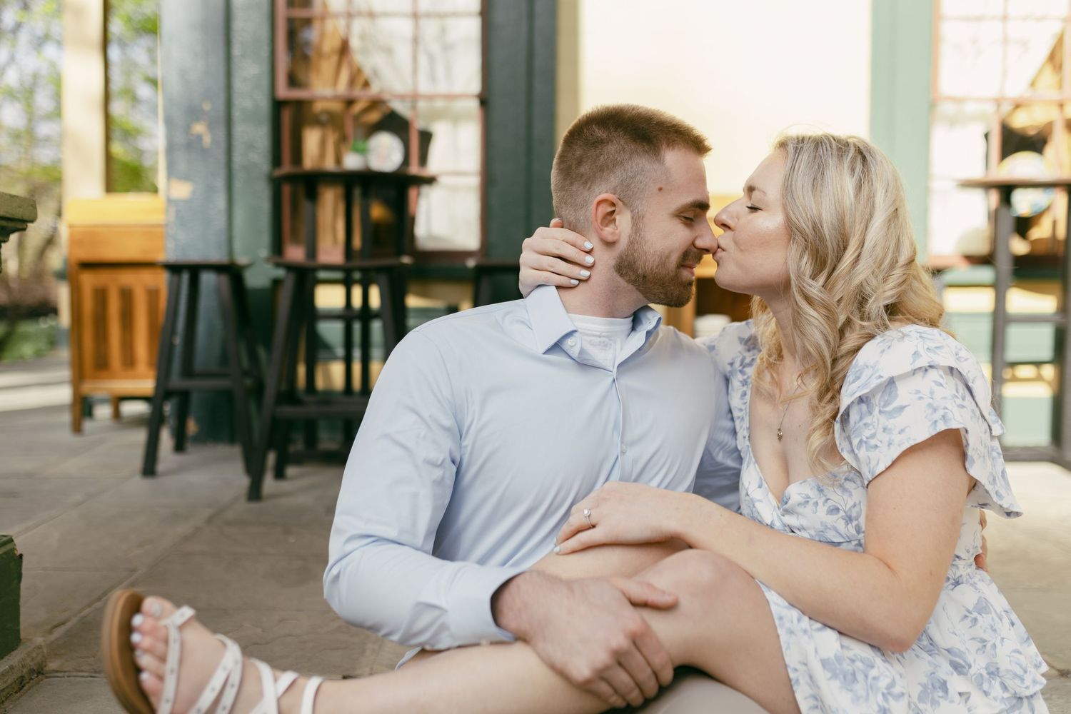 A smiling couple sitting closely together outside a cafe, the woman leaning in to kiss the man in a tender moment.