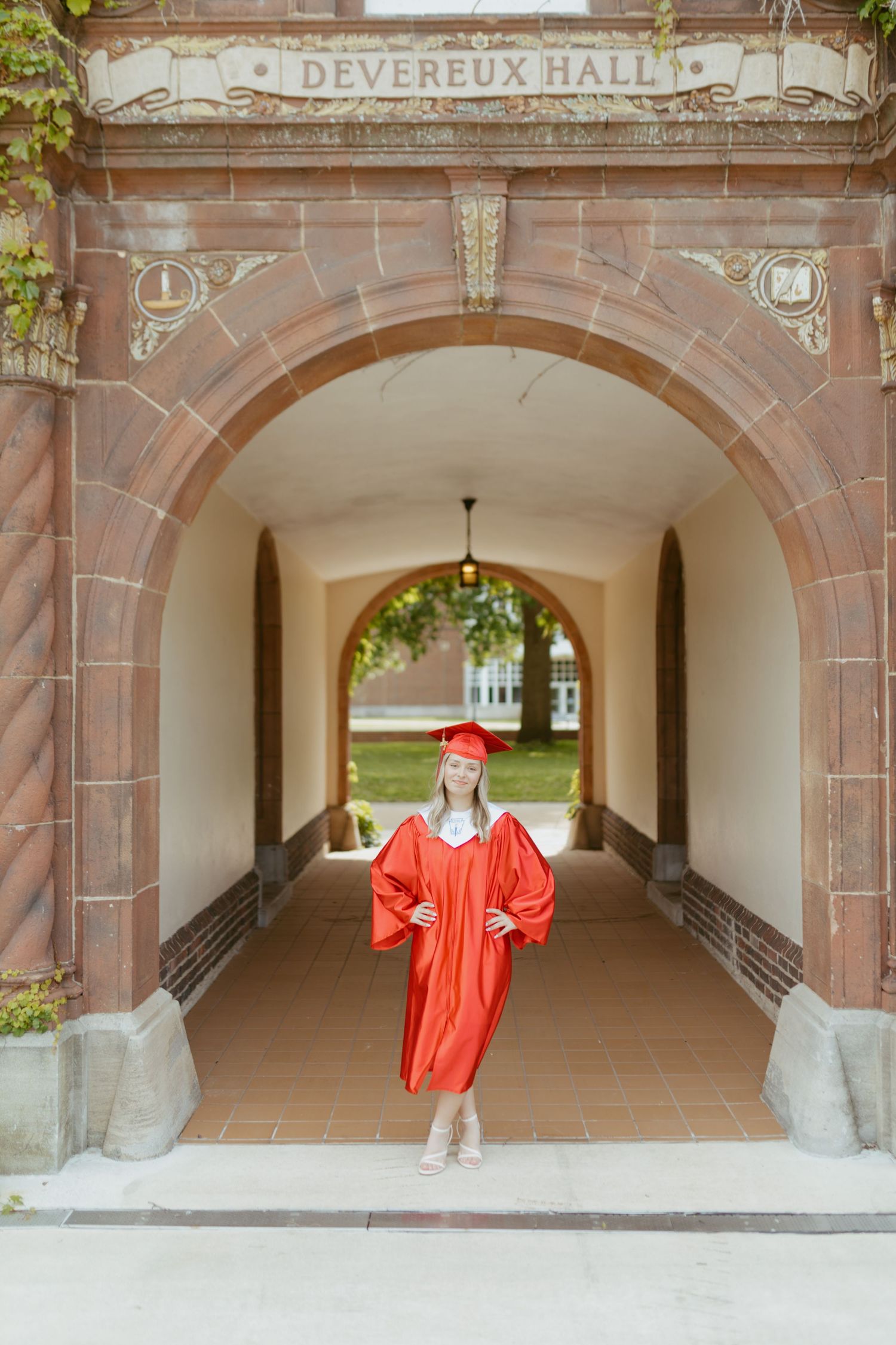Graduate in red cap and gown posing under Devereux Hall archway.