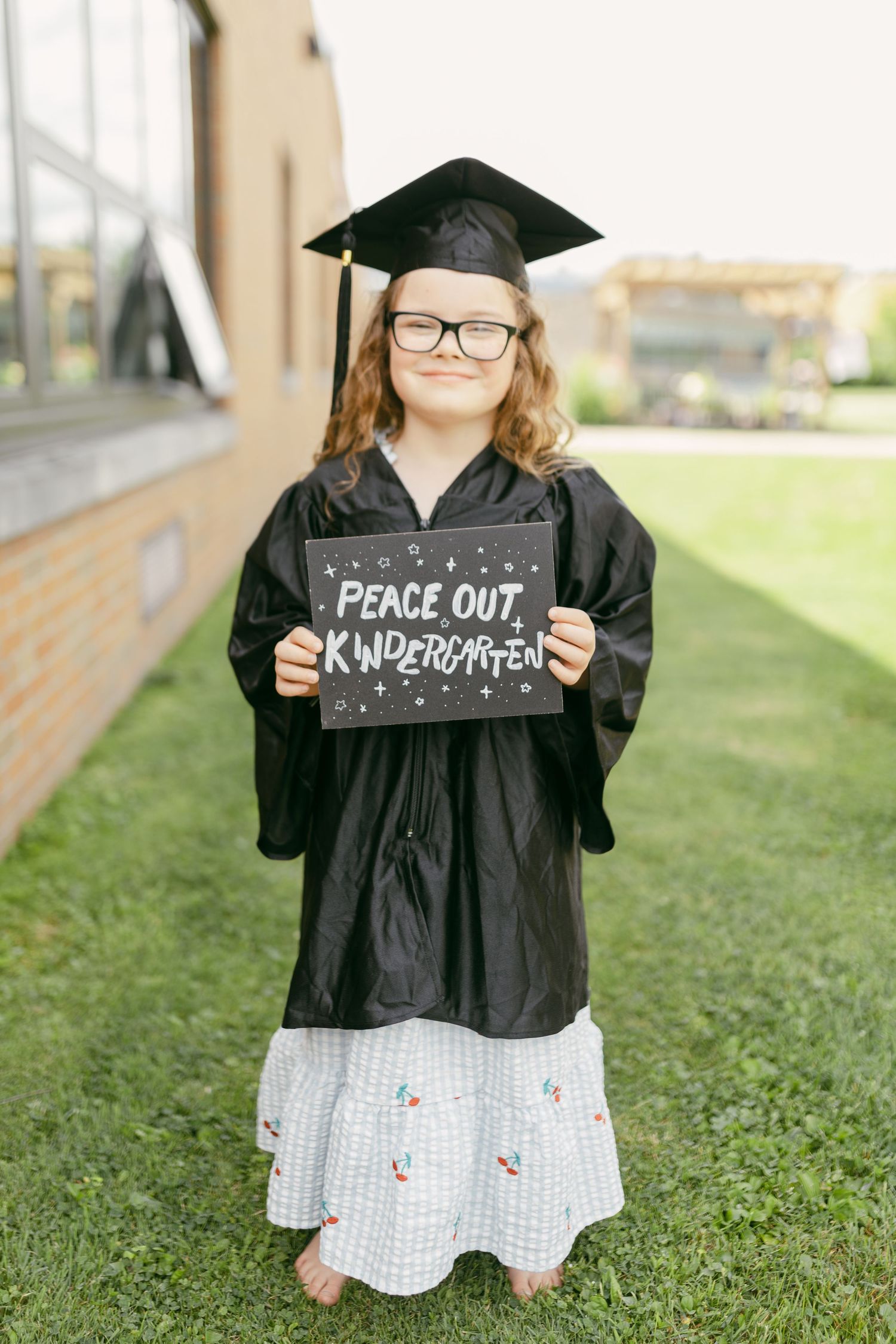 Young girl in graduation cap and gown holds a chalkboard sign reading Peace Out Kindergarten on green grass.