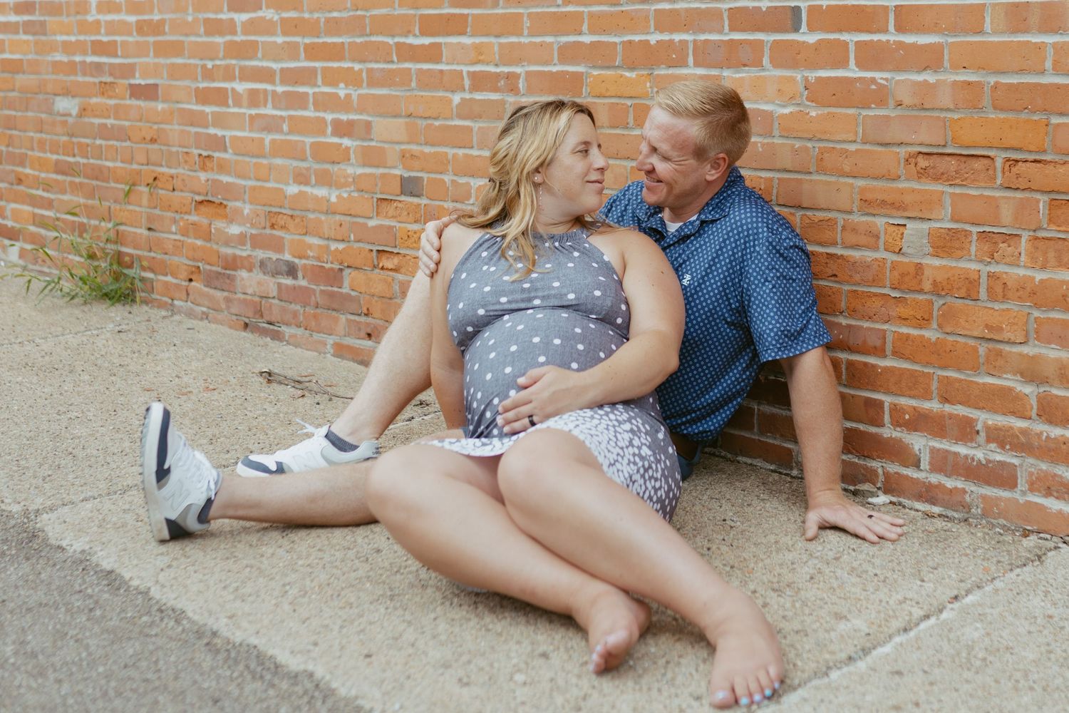 Pregnant woman and man sitting together against a brick wall, sharing a tender moment outdoors.