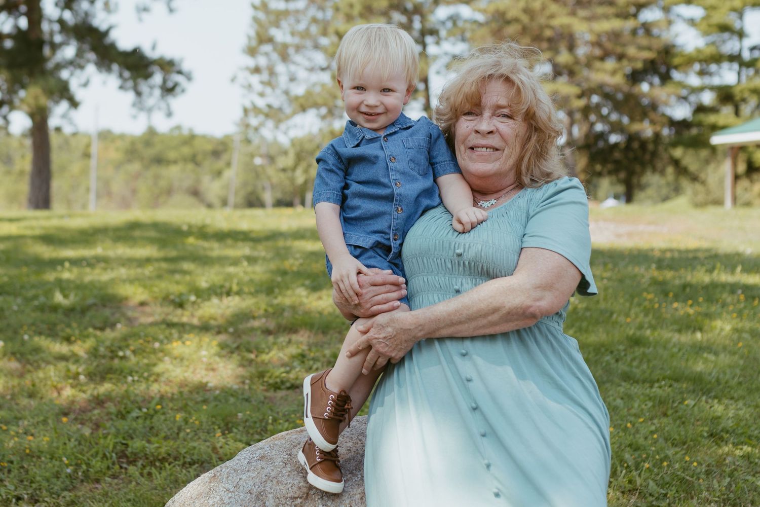 Grandmother sitting on a rock in a park, smiling while holding a young toddler boy in a blue denim outfit.