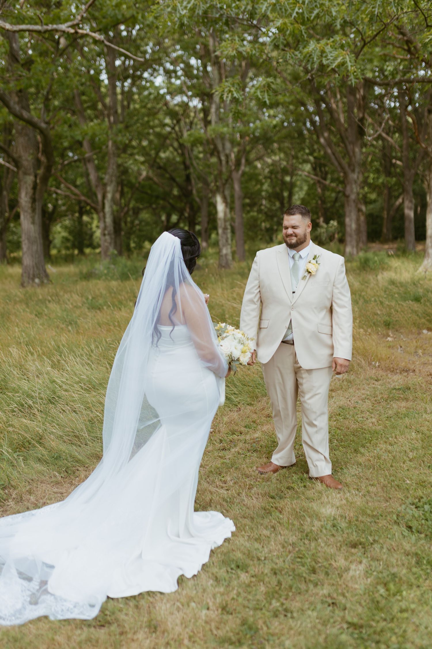 Bride in white gown with veil holds bouquet as groom in tan suit smiles during outdoor first look.