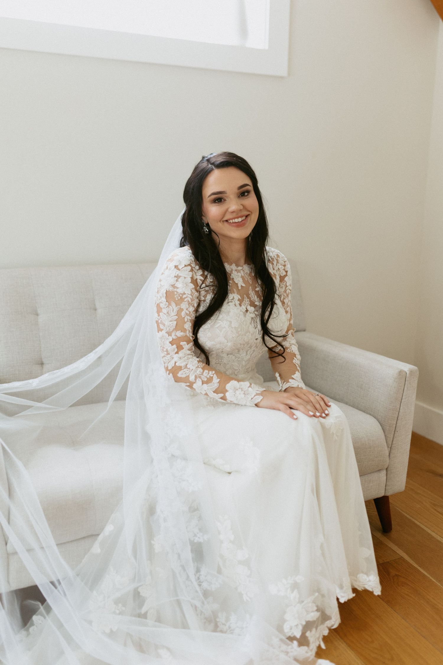 A bride sits elegantly on a white tufted chair wearing a detailed lace wedding gown with long sleeves.